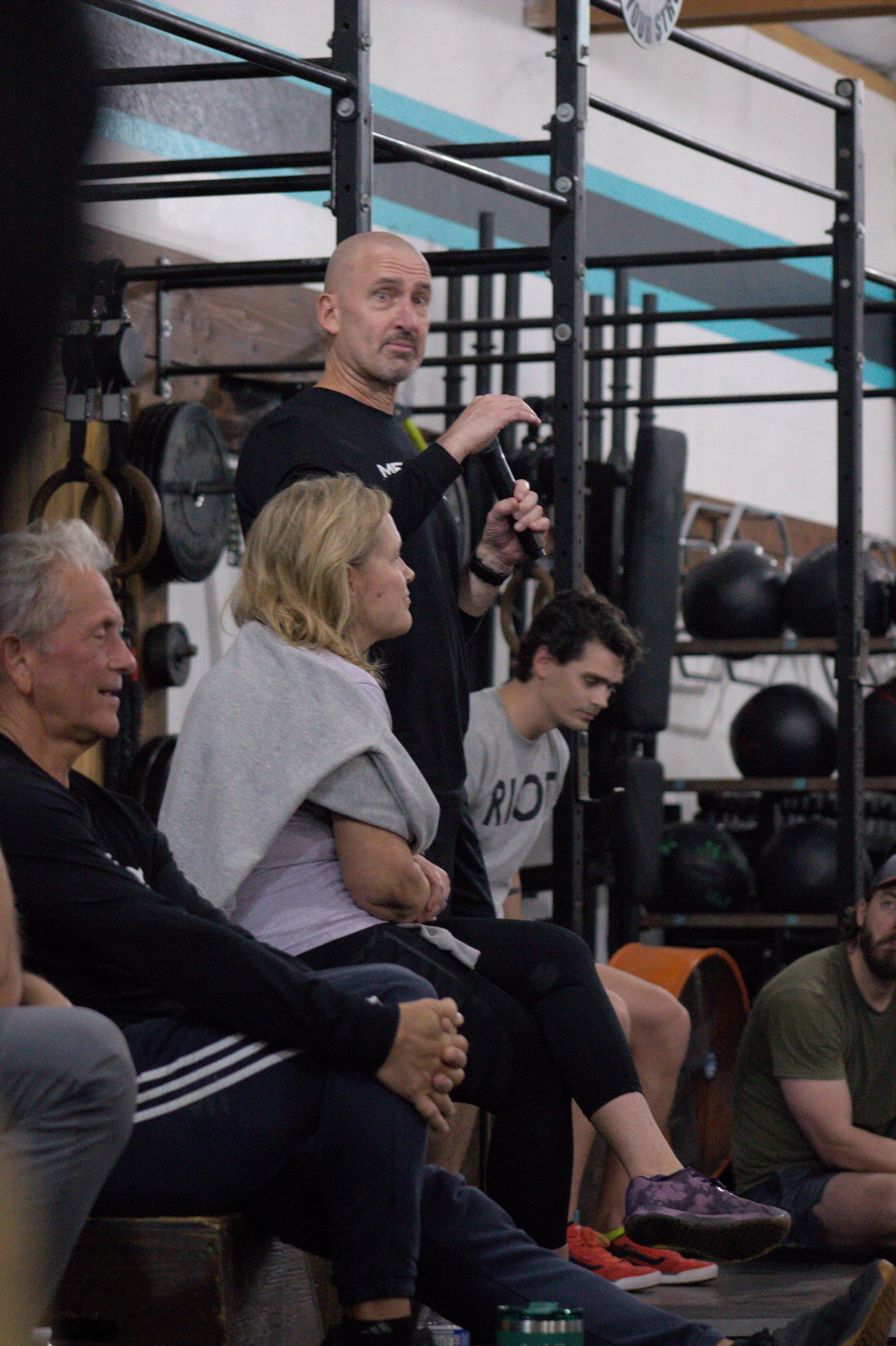 A man standing and speaking into a microphone in a gym with weights and gym equipment behind him, while three other people are seated and listening.