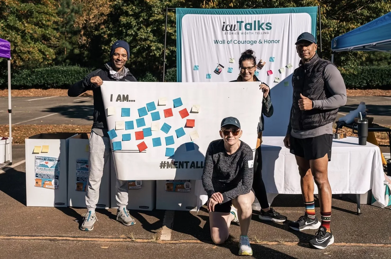 Group of five people participating in an outdoor mental health awareness event, holding a large sign with colorful post-it notes, with a banner behind them that reads 'icuTalks' and 'Wall of Courage & Honor.'