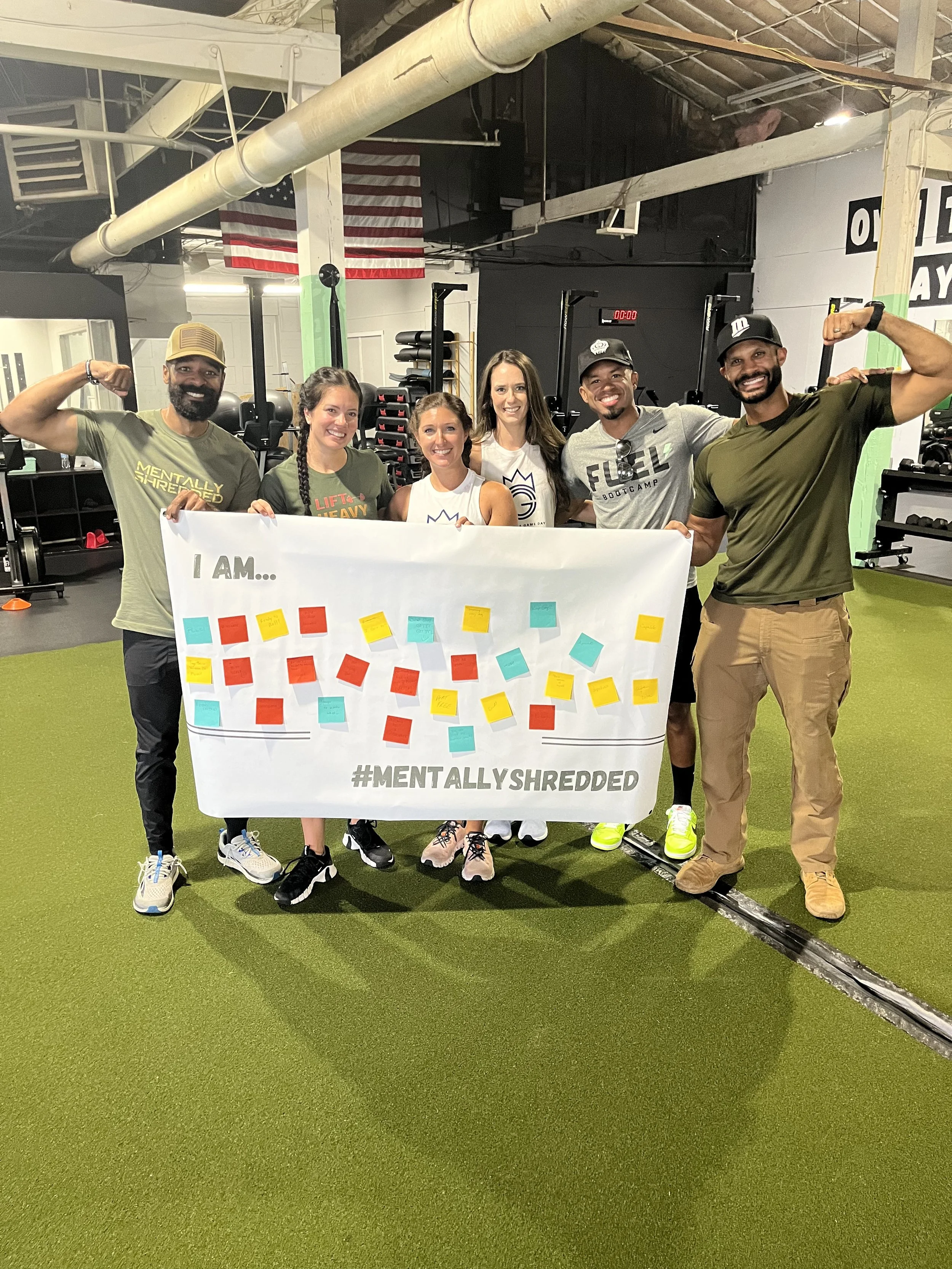 Group of six people in a gym holding a white banner with colorful sticky notes and the hashtag #MENTALLYSHREDDED. They are smiling, with some flexing muscles, and are dressed in workout attire.