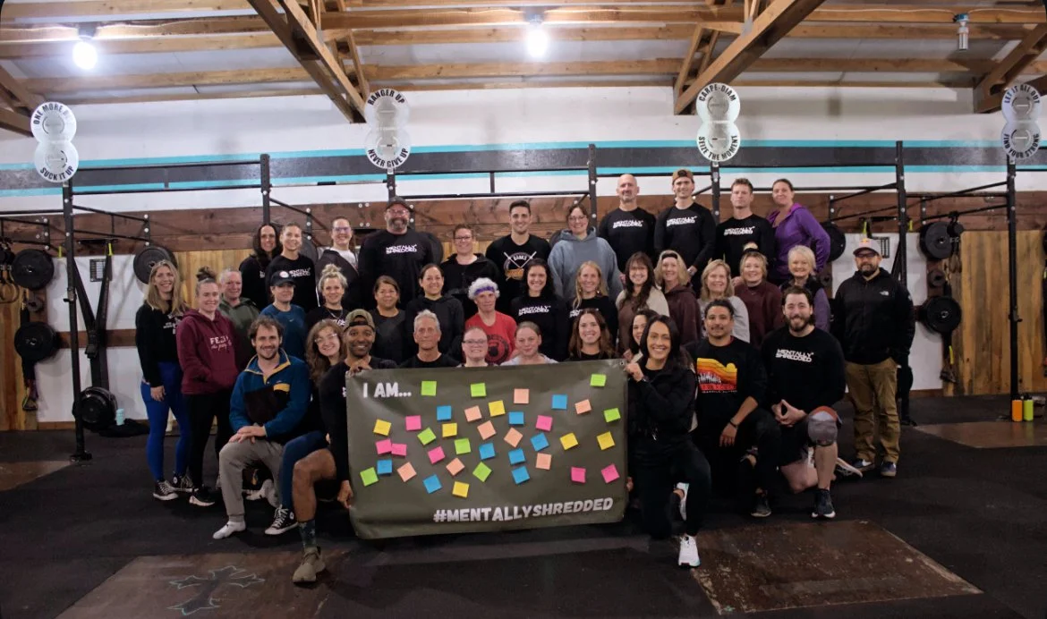 Group of people in a gym holding a sign with colorful sticky notes, gym equipment in the background, and wood-paneled walls.