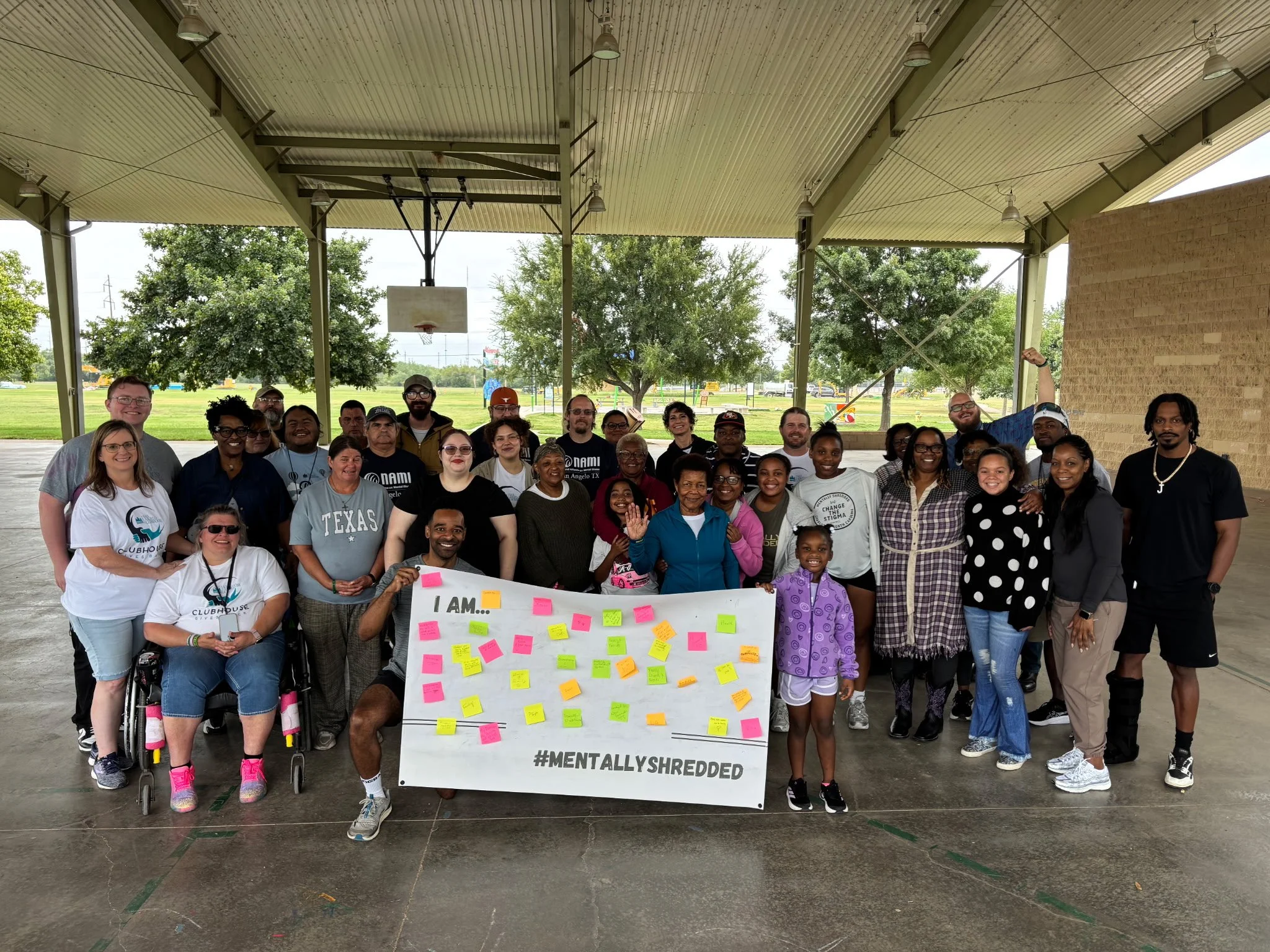 A diverse group of people posing under a pavilion with a basketball hoop in the background. They are holding a large white banner with colorful sticky notes and the hashtags #MENTALLYSHREDDED, and the words 'I AM...' written at the top.