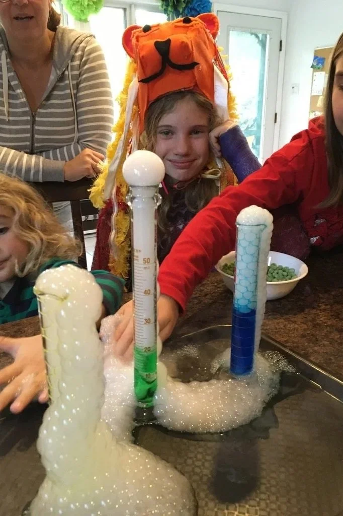 Enfants en train de faire une expérience scientifique avec des tubes à essai dans une cuisine, un enfant porte un chapeau en forme de tigre.