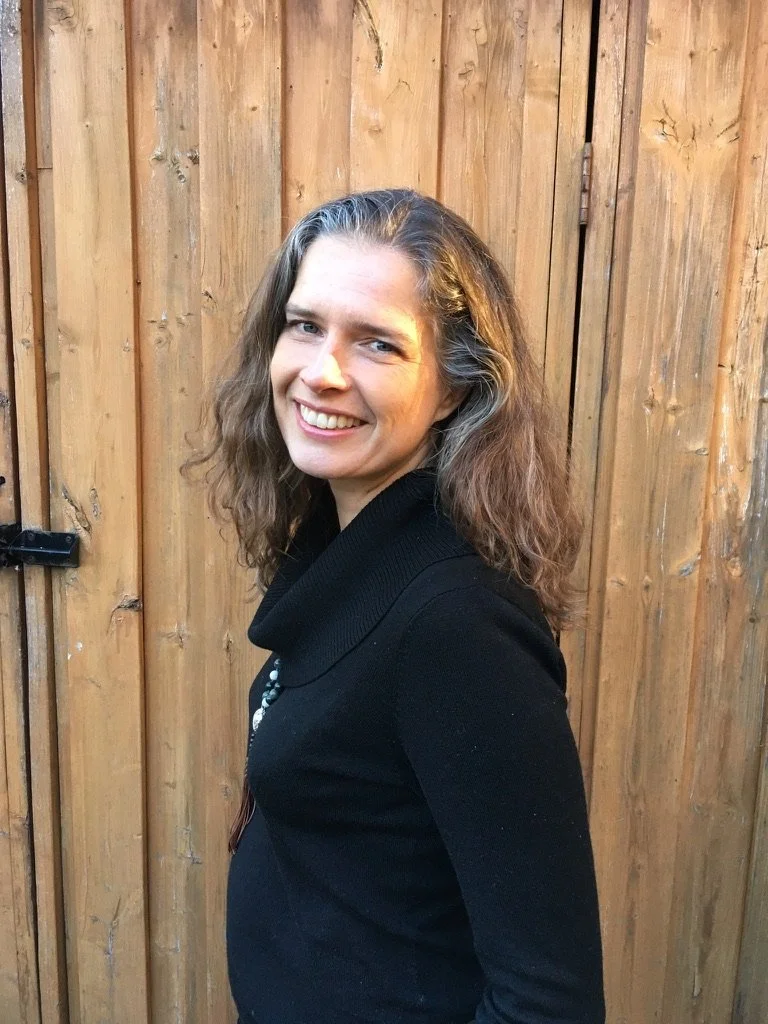 A woman with long wavy hair, smiling, wearing a black top, standing in front of a wooden fence.