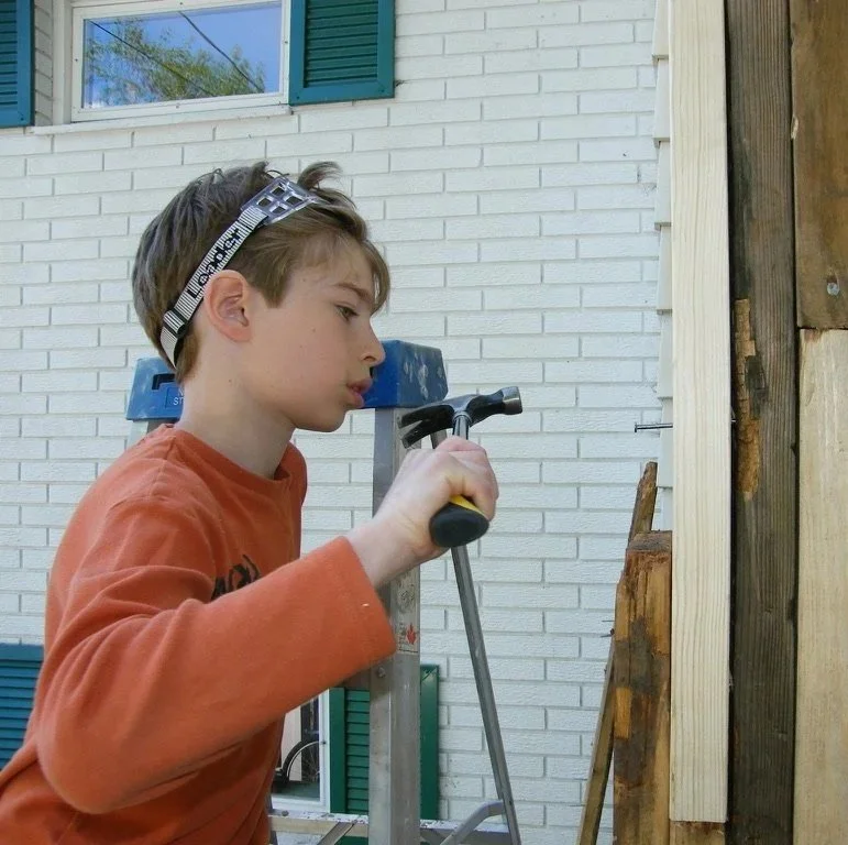 Un garçon avec des lunettes de protection utilise un marteau pour travailler sur une structure en bois à l'extérieur, devant une maison avec des murs en briques blanches et des volets verts.