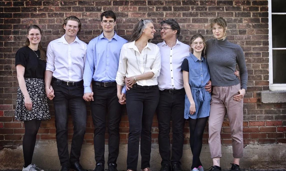 Seven family members standing against a brick wall, holding hands, smiling, and looking at each other.