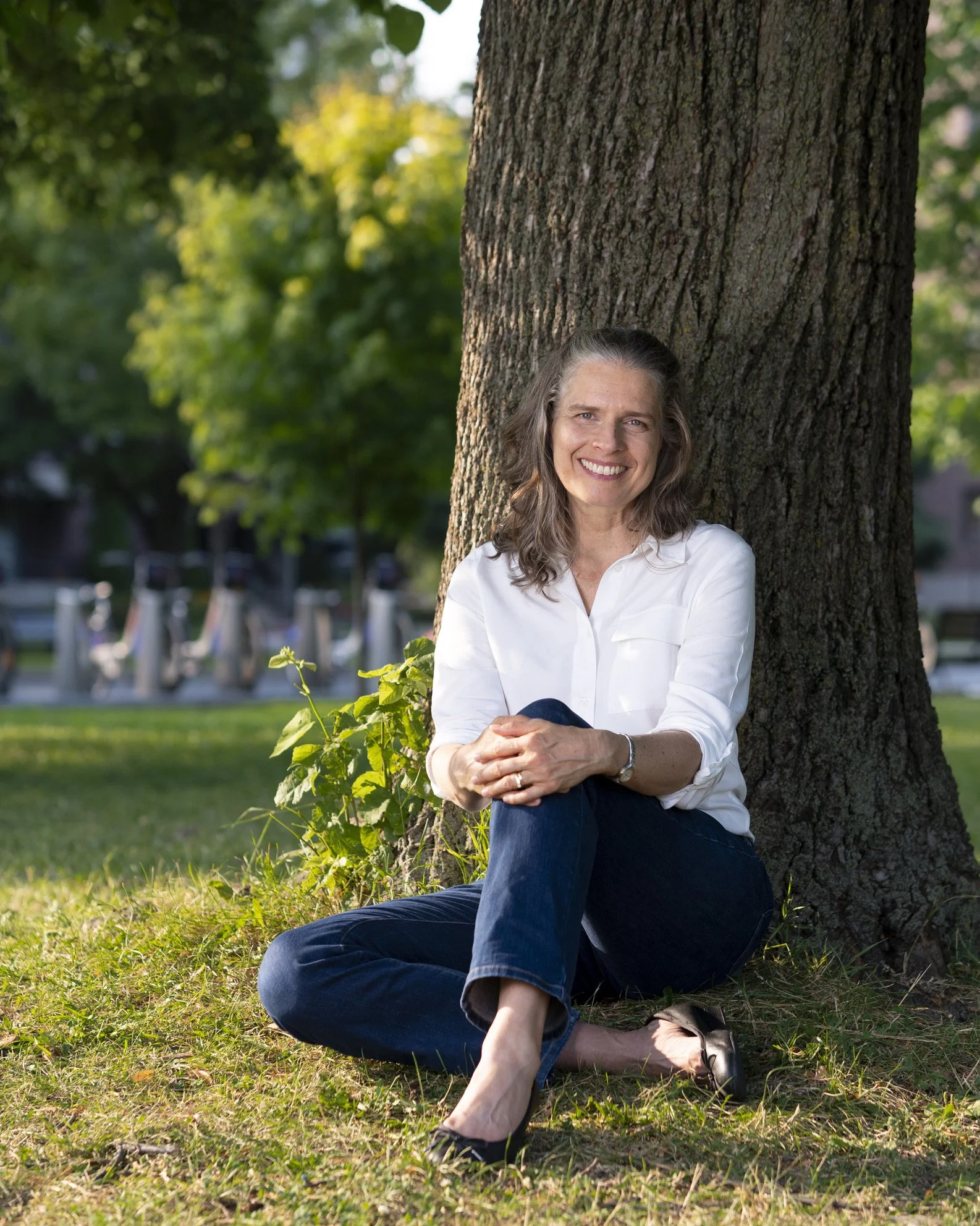 A woman with gray hair wearing a white shirt and dark jeans sitting on the grass next to a tree in a park, smiling at the camera.