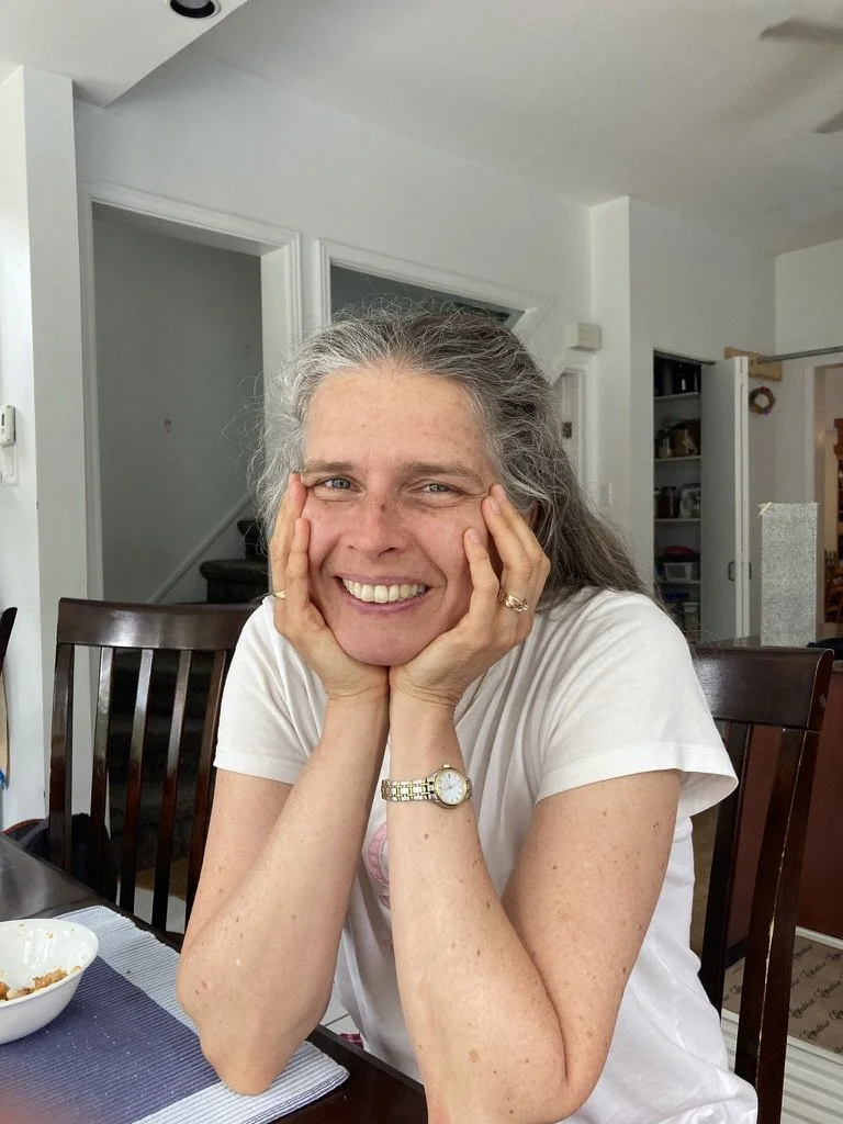 A woman with gray hair smiling and resting her face in her hands at a dining table in a home kitchen.