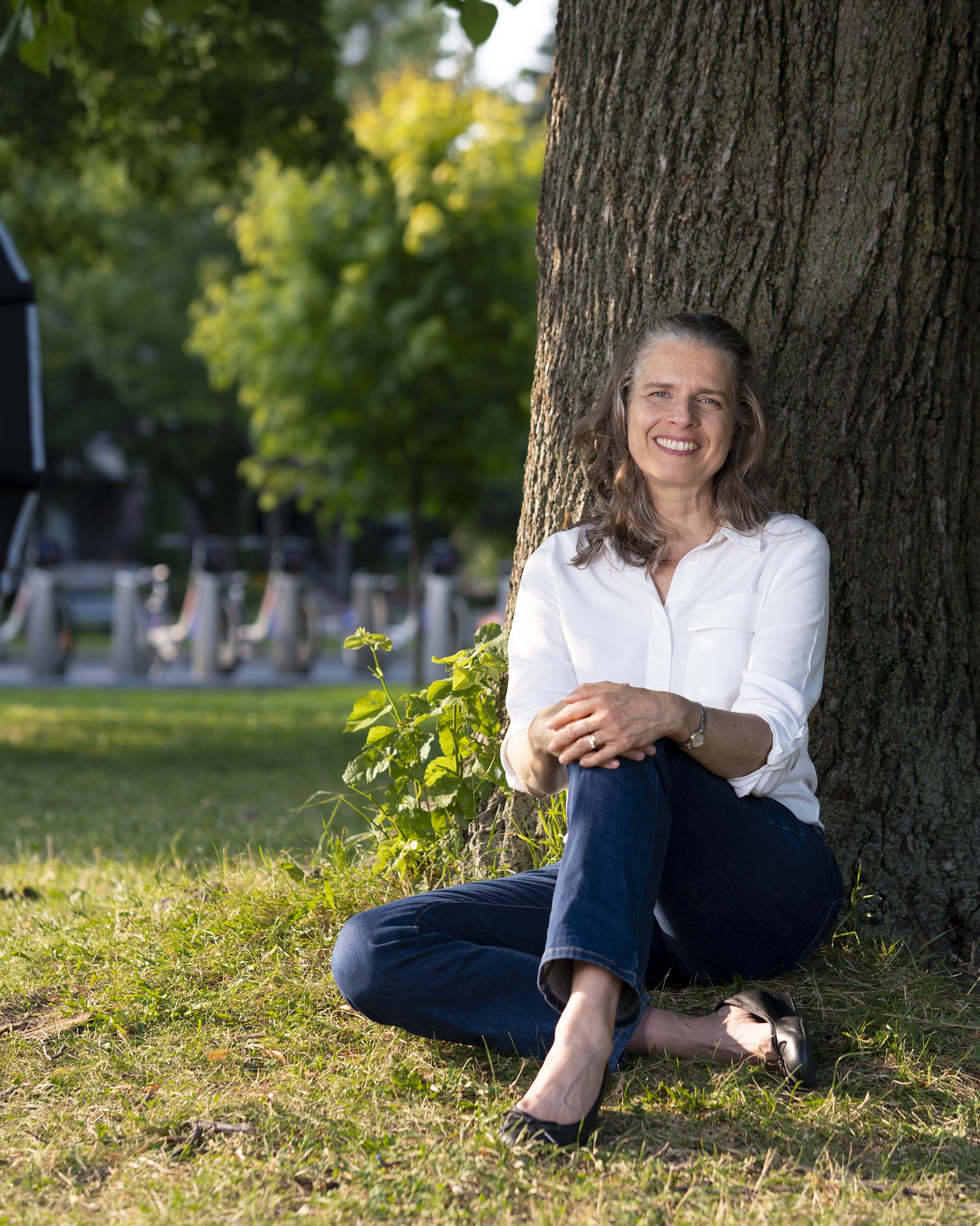 Une femme assise contre un arbre dans un parc, souriant, avec des cheveux gris, portant une chemise blanche et un pantalon en jean.