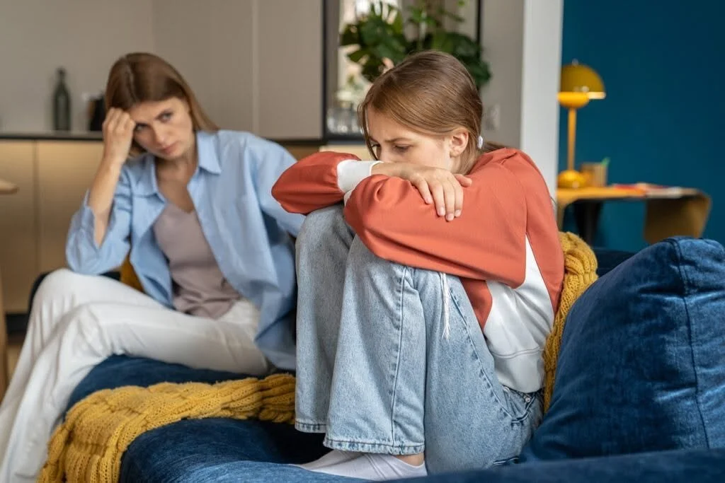 Une femme semble triste ou préoccupée, assise sur un canapé avec une jeune fille qui pleure ou est en détresse, dans un intérieur moderne.