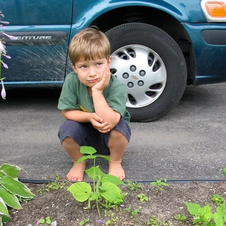 Un garçon assis par terre devant une voiture, regardant vers l'objectif, dans un jardin avec des plantes.