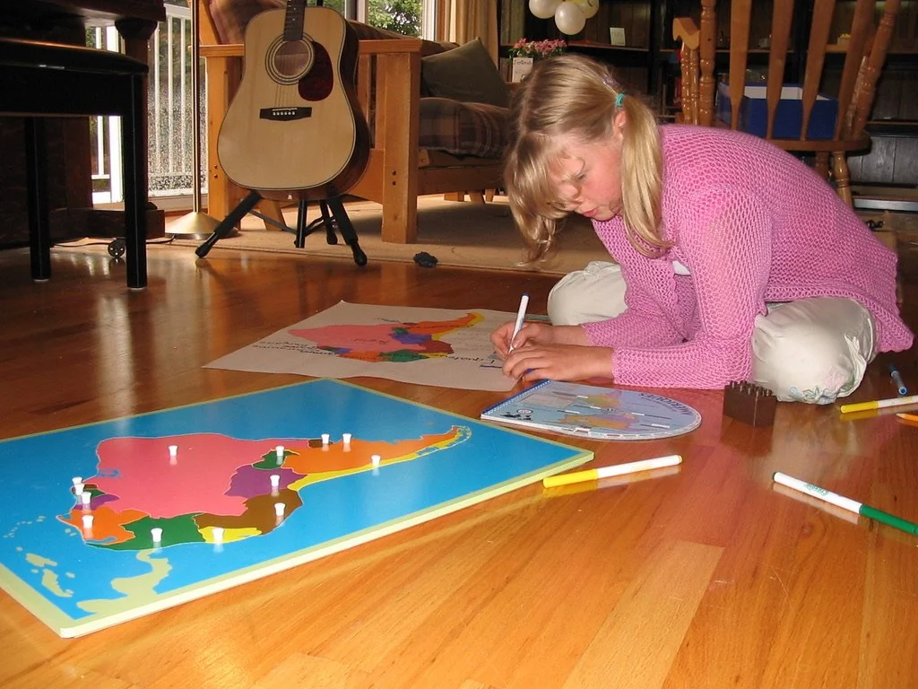 Une jeune fille assise sur le sol joue à un jeu de société avec une carte et des figurines représentant une carte du Canada, à côté d'une guitare dans un salon.
