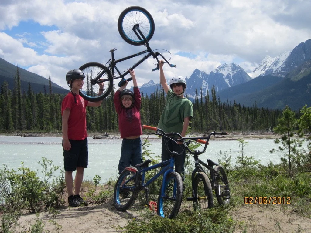 Three children by a river with mountains in the background, holding a bicycle in the air, with two additional bikes on the ground.