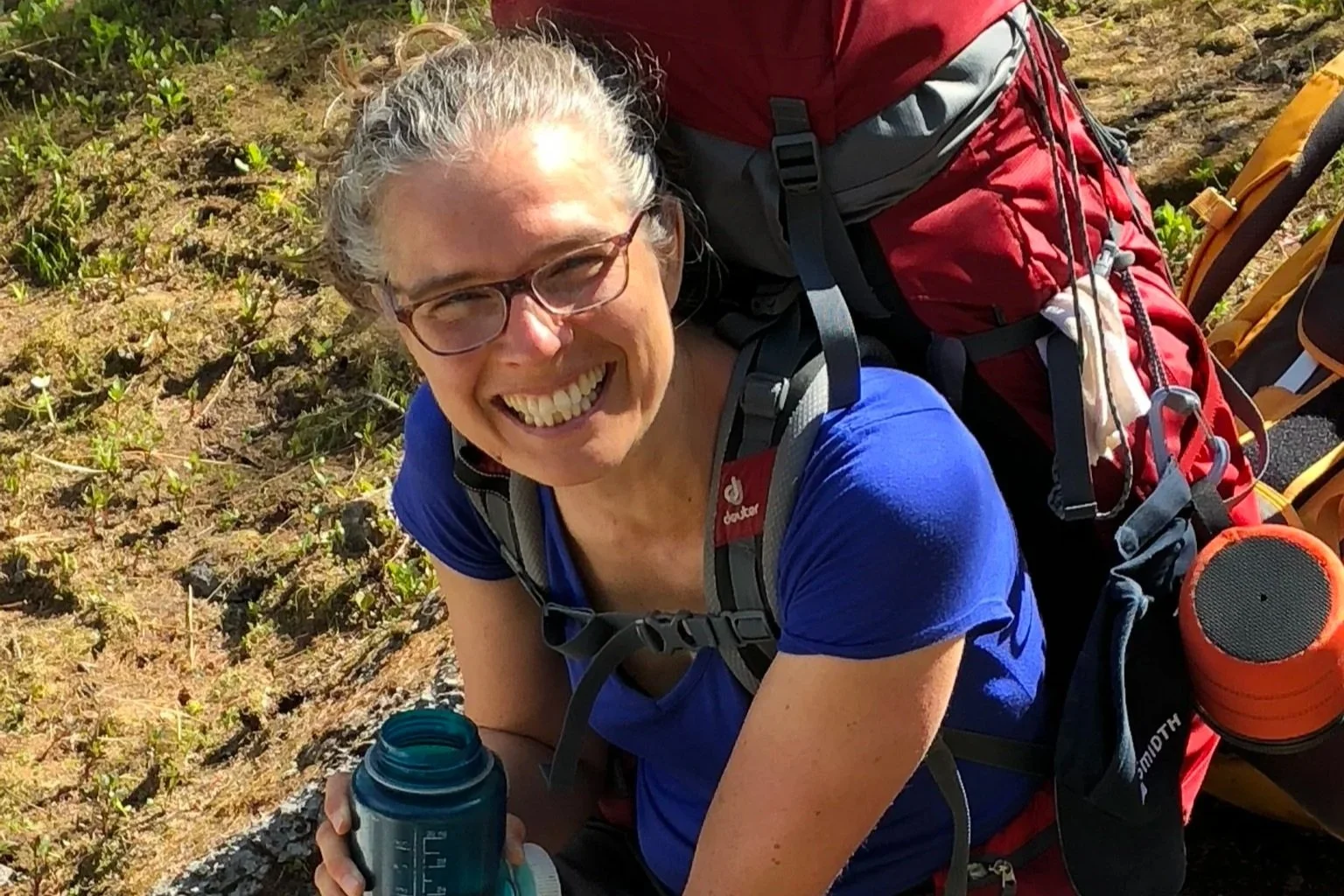 Une femme souriante portant des lunettes, avec un sac à dos rouge, en pleine randonnée en plein air, tenant une bouteille d'eau. Elle porte un t-shirt bleu et semble heureuse.
