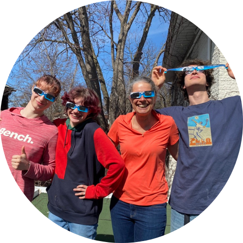 Quatre personnes portent des lunettes de protection de solar et sourient sous un ciel bleu, avec des arbres sans feuilles en arrière-plan.