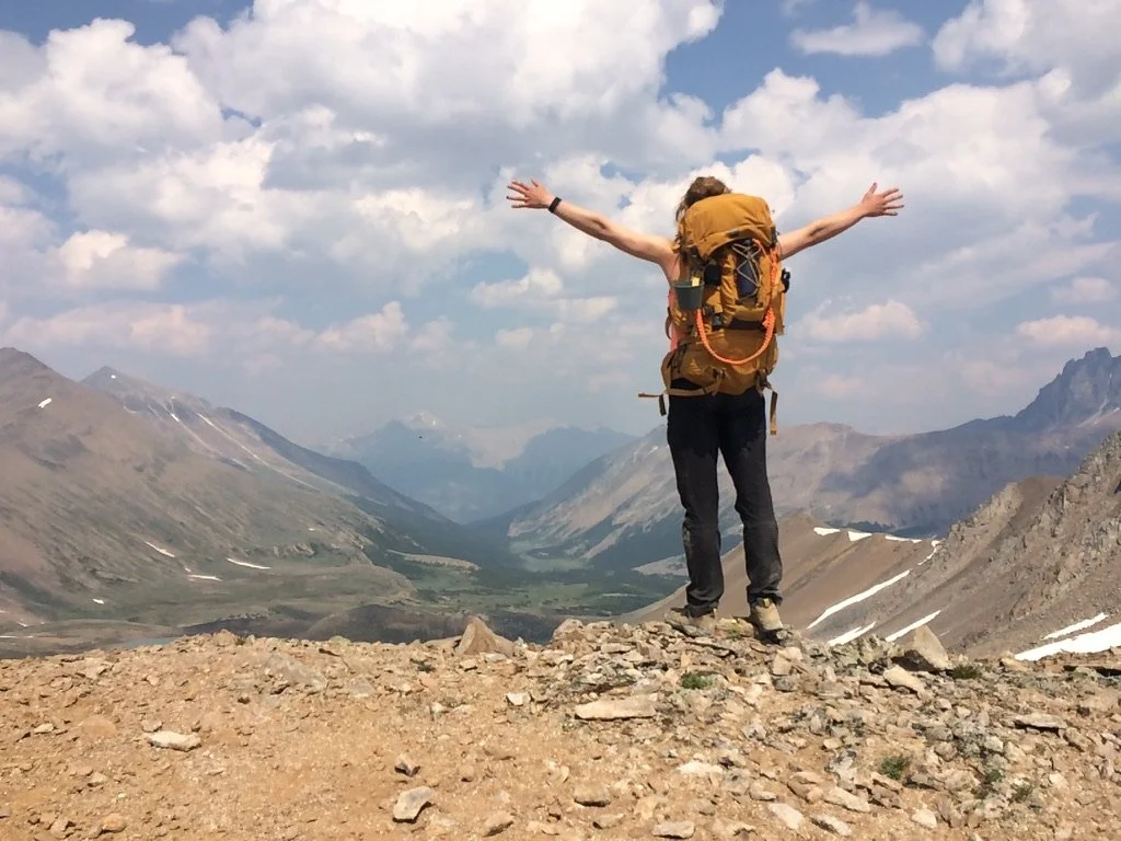Une personne avec un sac à dos grand exposée sur une montagne, avec un paysage de montagnes et de vallées en arrière-plan, sous un ciel nuageux.