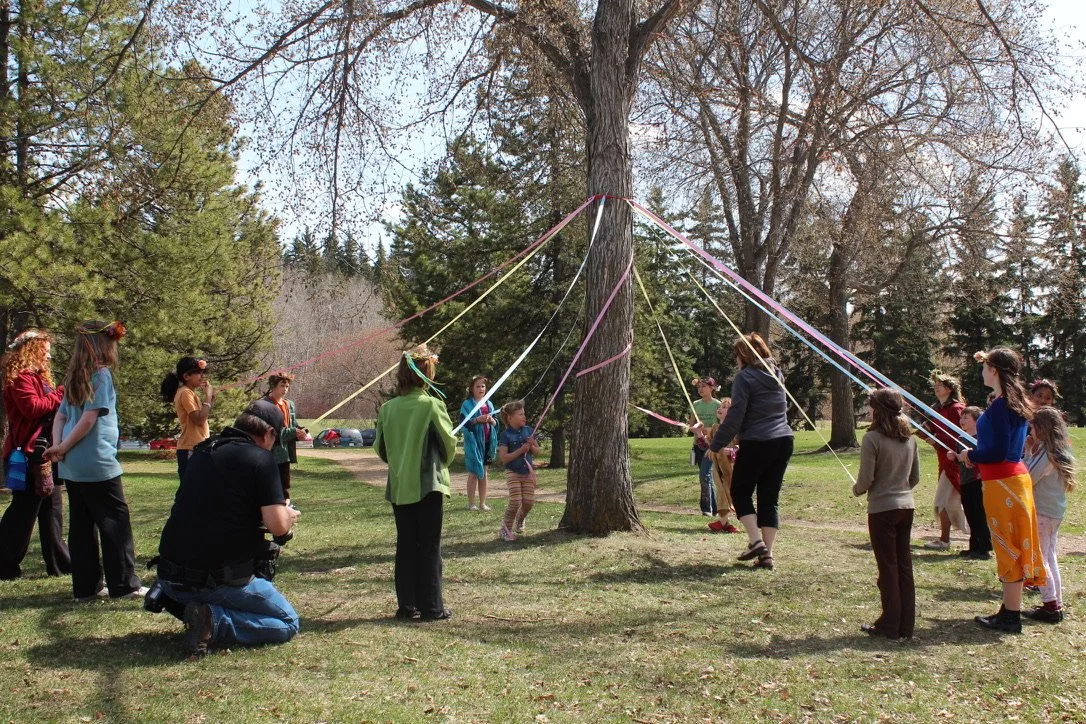 Groupe de personnes, adultes et enfants, participant à une activité de jeu ou d'équipe autour d'un arbre, avec des rubans colorés attachés à l'arbre et tenu par les participants.