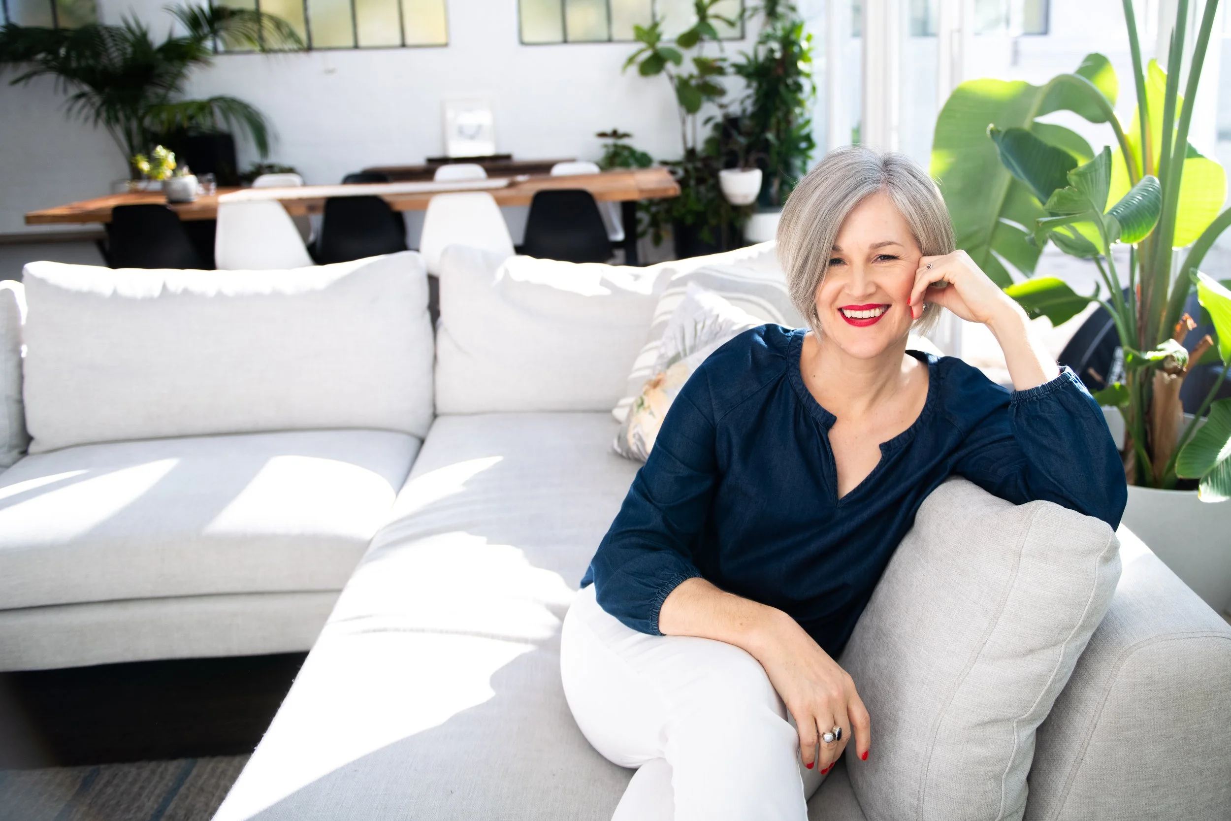 Anna sitting on a sofa in a sunny room with green foliage in the background. You are invited to join Anna in The Reading Room.