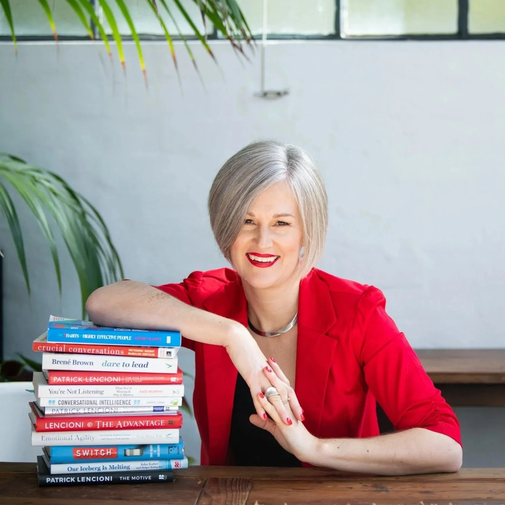 A woman with short gray hair, wearing a red blazer, smiling with red lipstick, sitting at a table with a stack of books in front of her. The books include titles like 'Crucial Conversations,' 'Dare to Lead,' and 'The Motive.' There are green plants and a window with natural light in the background.