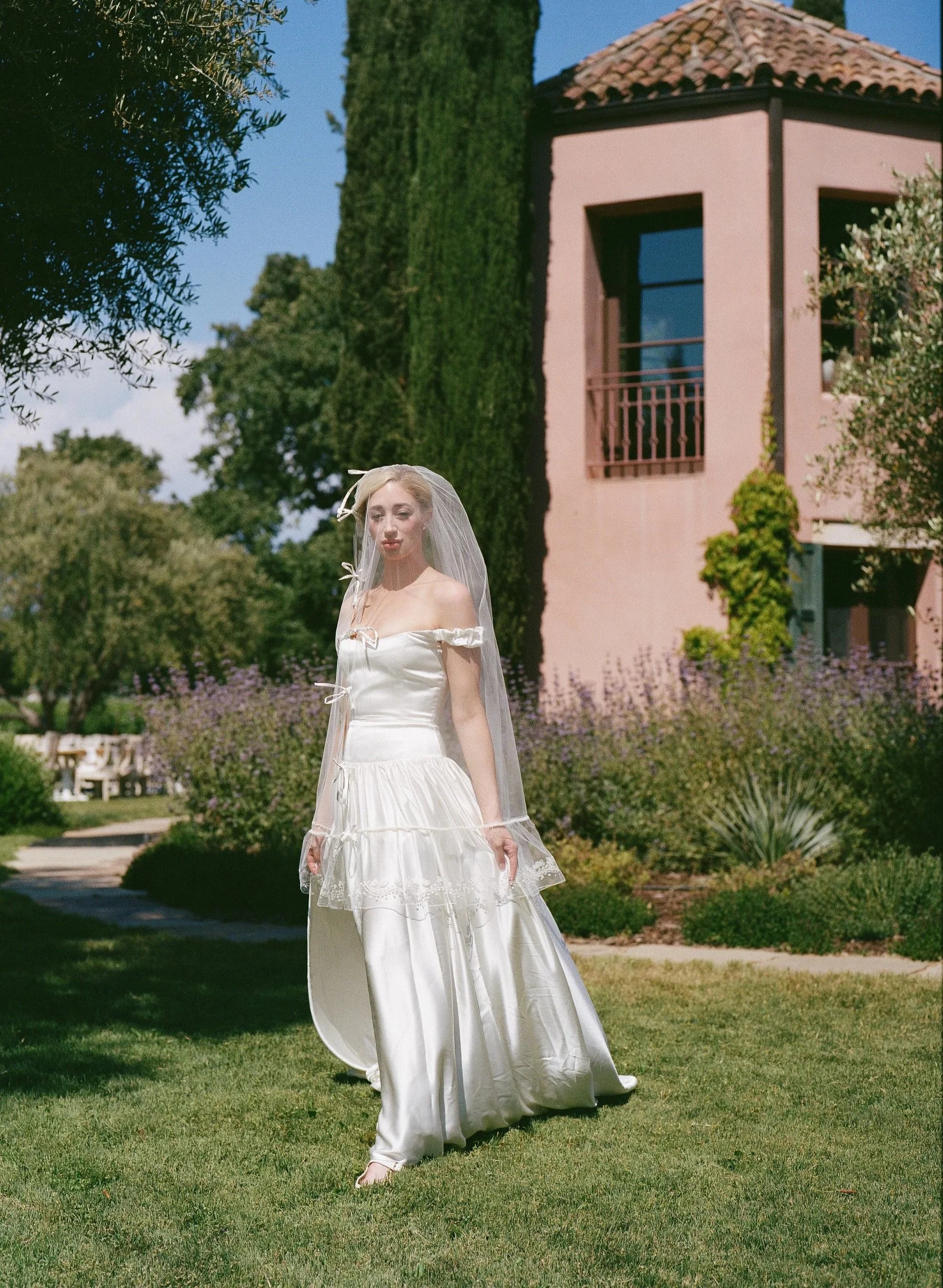 A woman in a white off-shoulder wedding dress and veil standing on a lawn in front of a pink building with a tile roof, surrounded by greenery and trees.