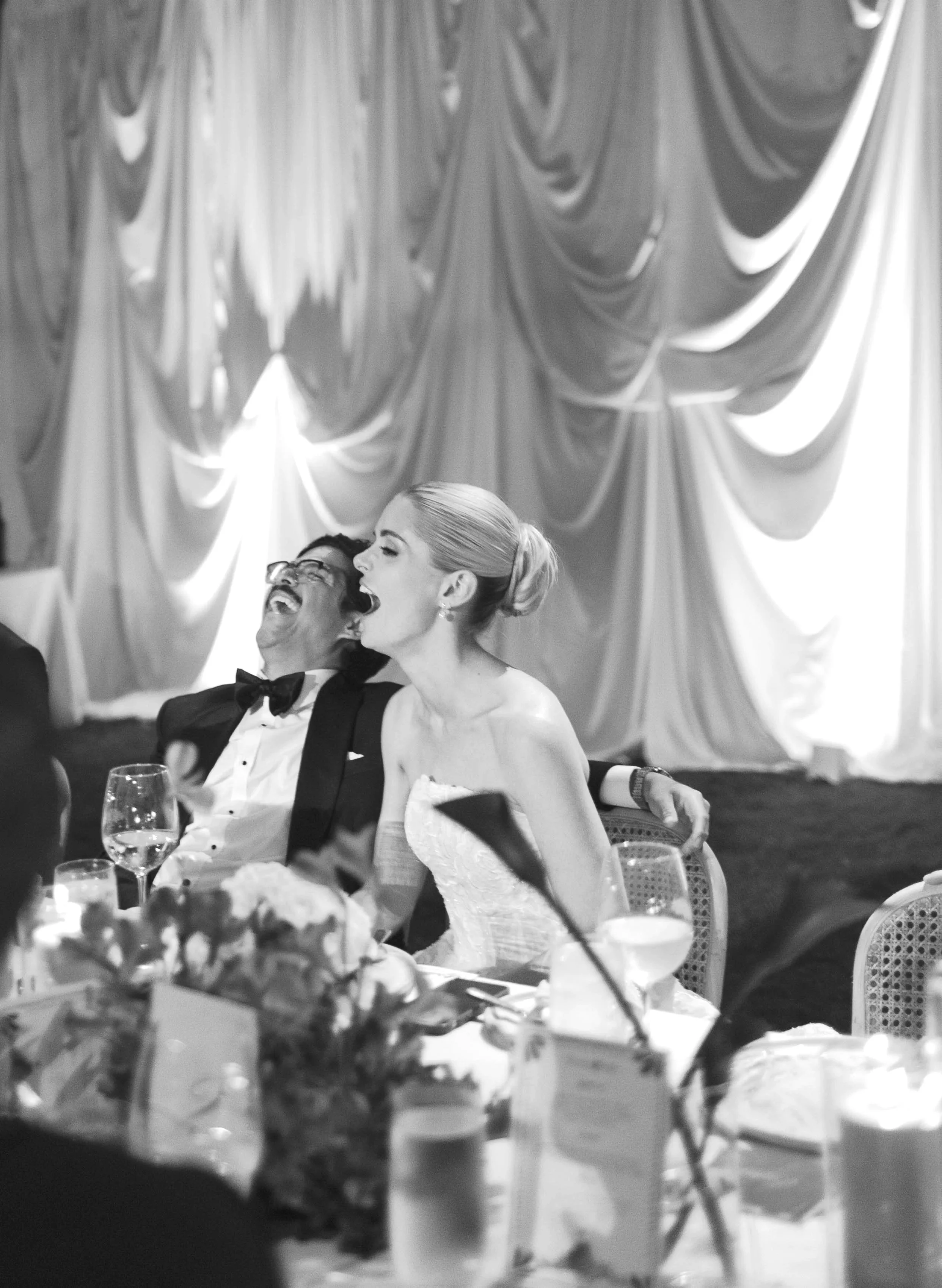 Black and white photo of a couple at a wedding reception, laughing and smiling together. The man is wearing a tuxedo with a bow tie and glasses, and the woman is in a strapless wedding gown with her hair in an elegant updo. They are seated at a table