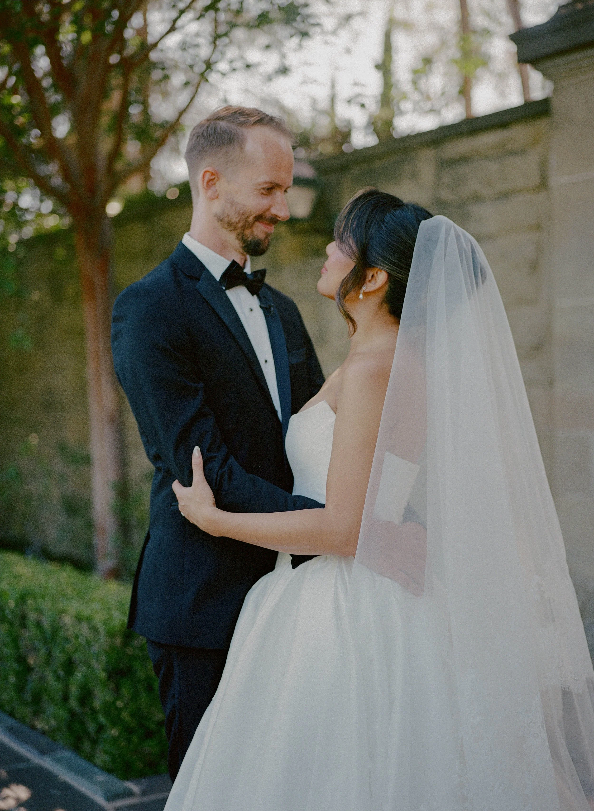 A bride and groom stand outdoors in formal wedding attire, gazing lovingly at each other, with a stone wall and trees in the background.