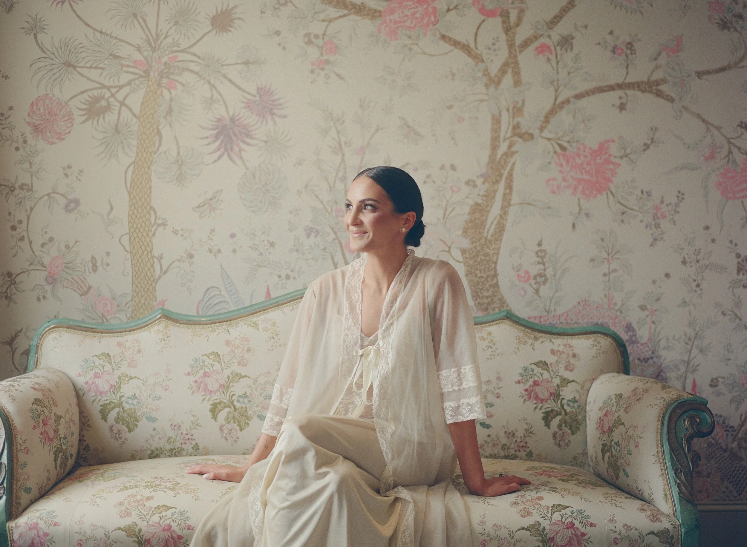 A woman in vintage cream-colored pajamas sitting on an ornate floral sofa in front of a wallpaper with nature and floral motifs.