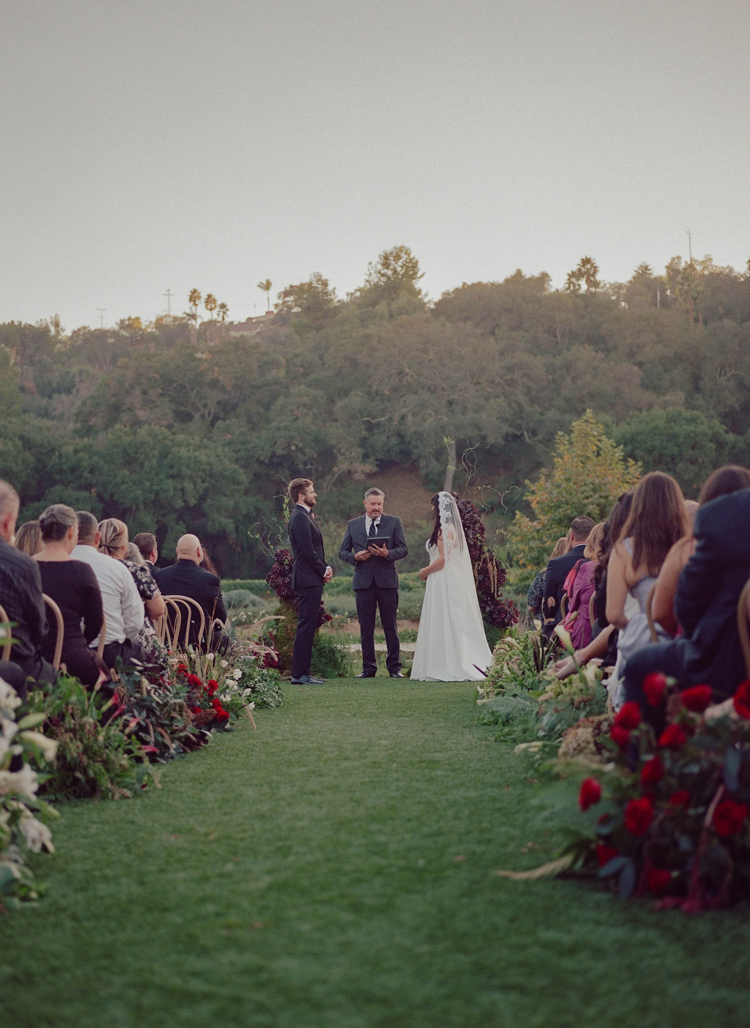 A wedding ceremony taking place outdoors on a grassy aisle with guests seated on both sides, a bride and groom facing each other, and a person officiating in the middle near the lake and trees backdrop.