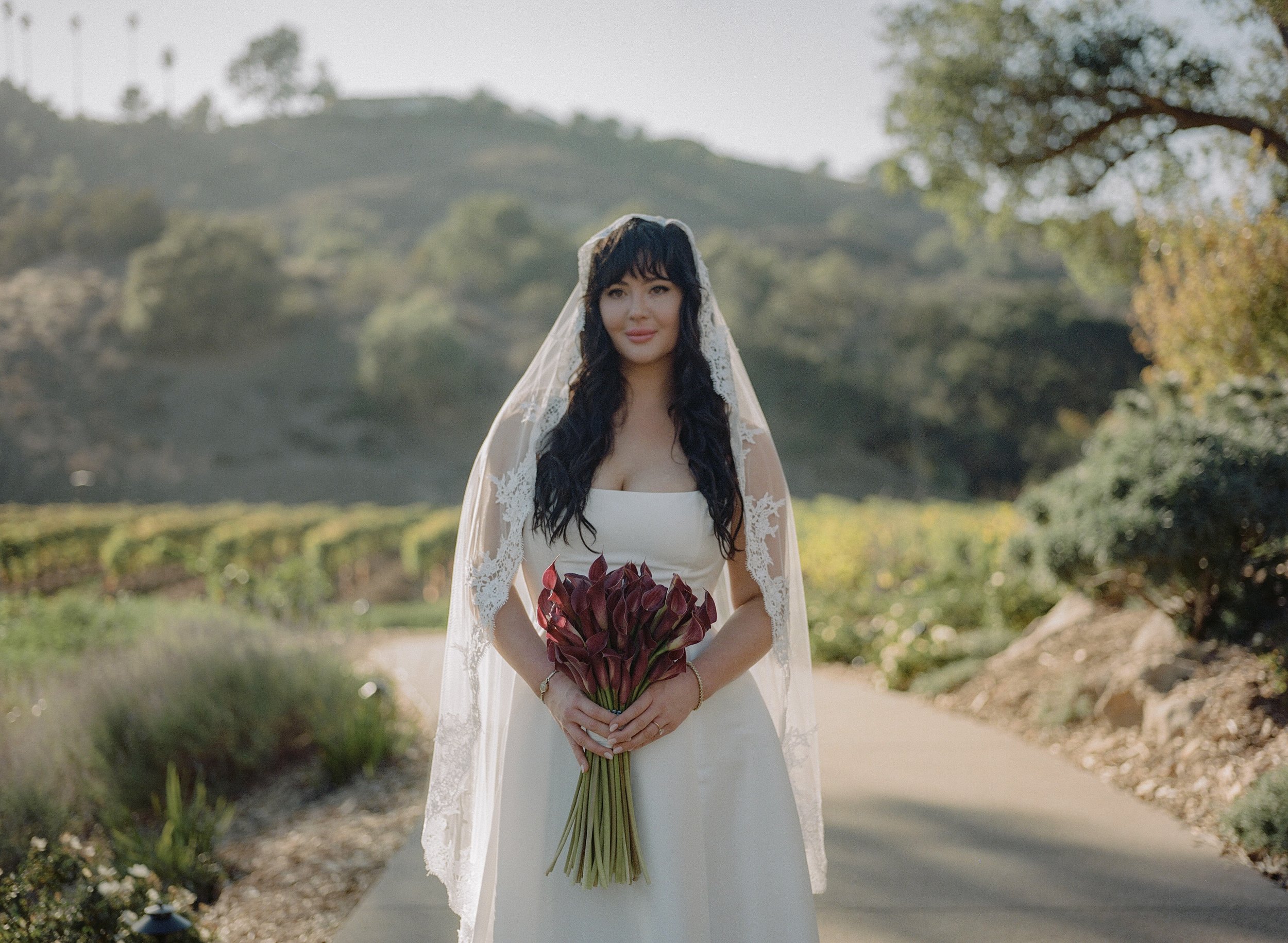 A woman in a white wedding dress, holding a bouquet of dark red calla lilies, standing outdoors on a paved path with greenery and hills in the background.