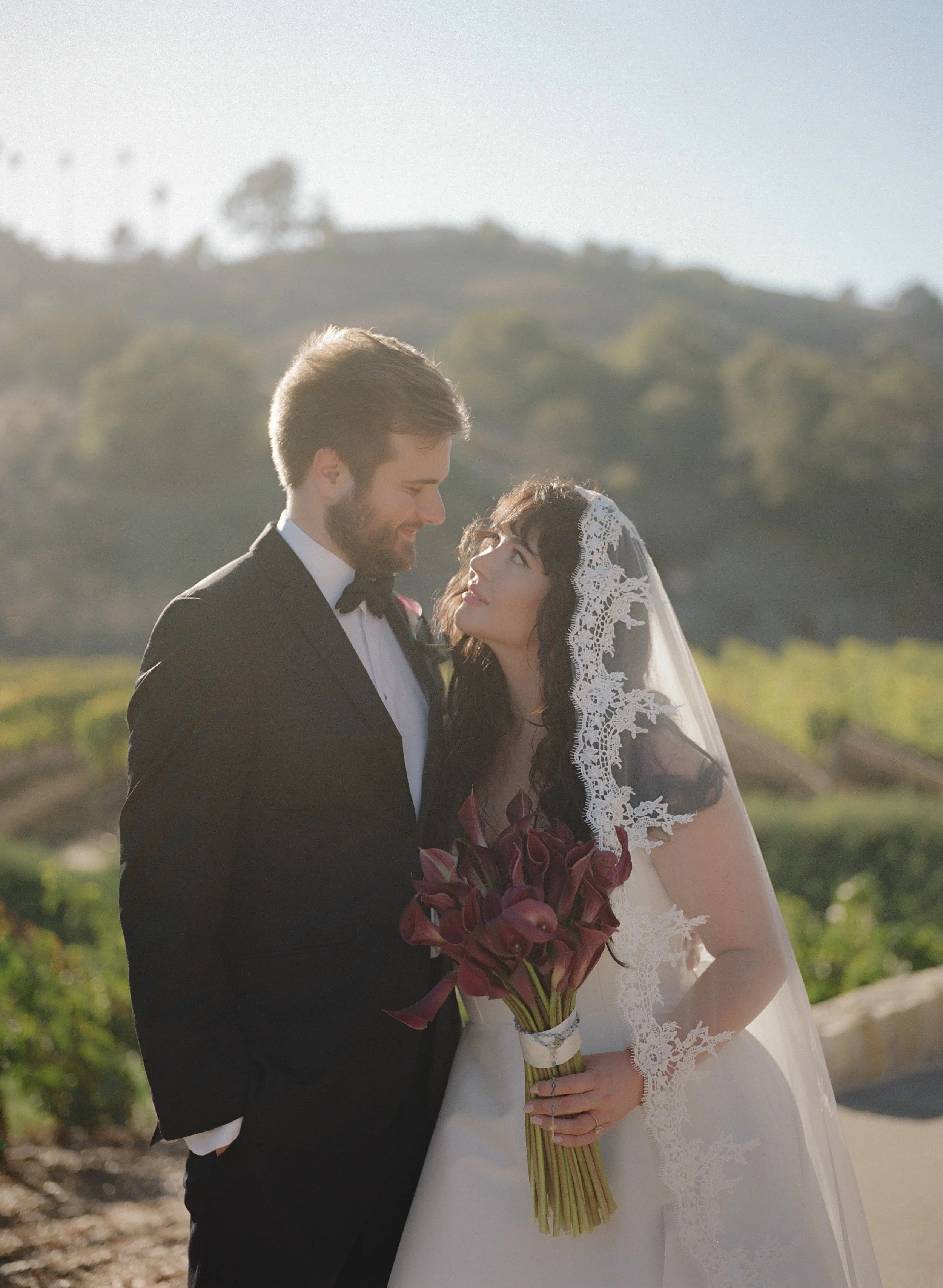 A bride and groom dressed in wedding attire standing outdoors, gazing into each other's eyes, with a scenic landscape of hills and greenery in the background. The bride holds a bouquet of dark red calla lilies and wears a lace veil.