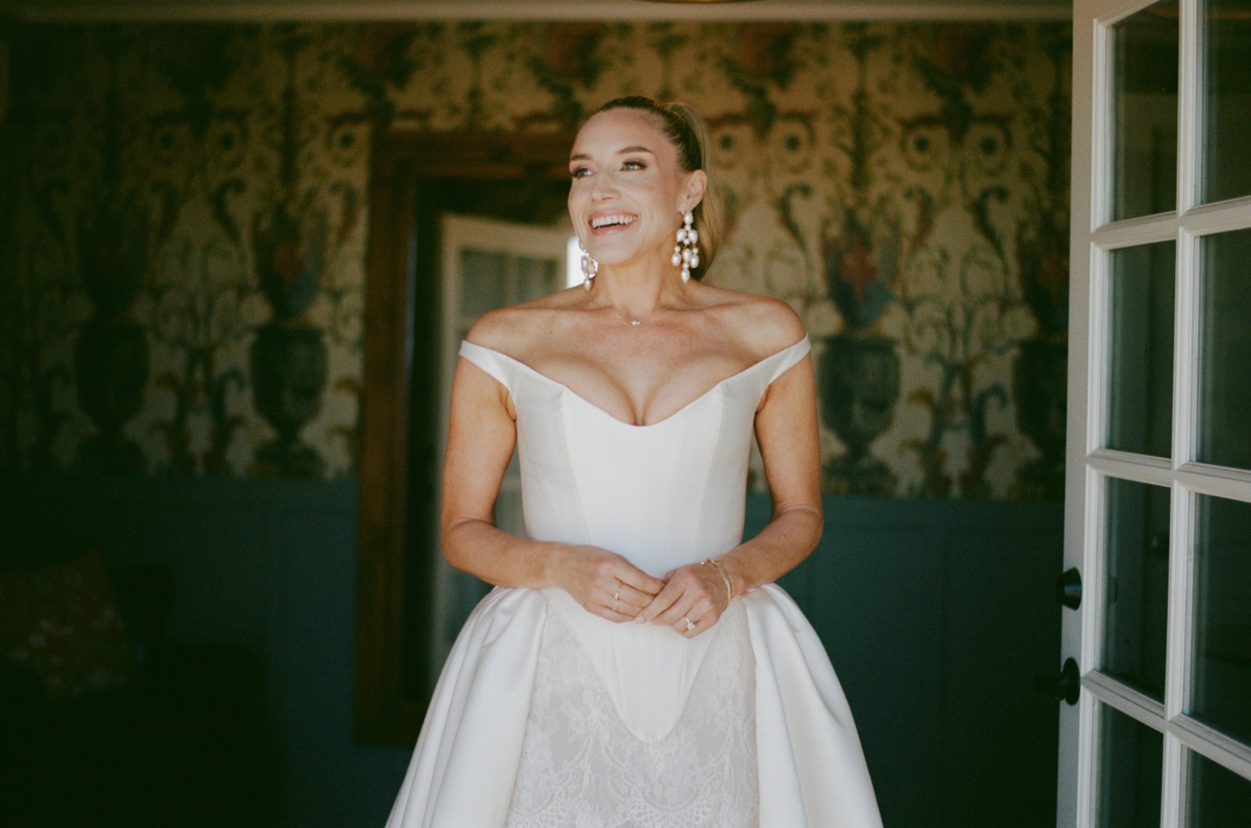 A woman in a wedding dress with off-the-shoulder design, pearl earrings, and makeup, standing in a room with ornate wallpaper, smiling and looking to her left.