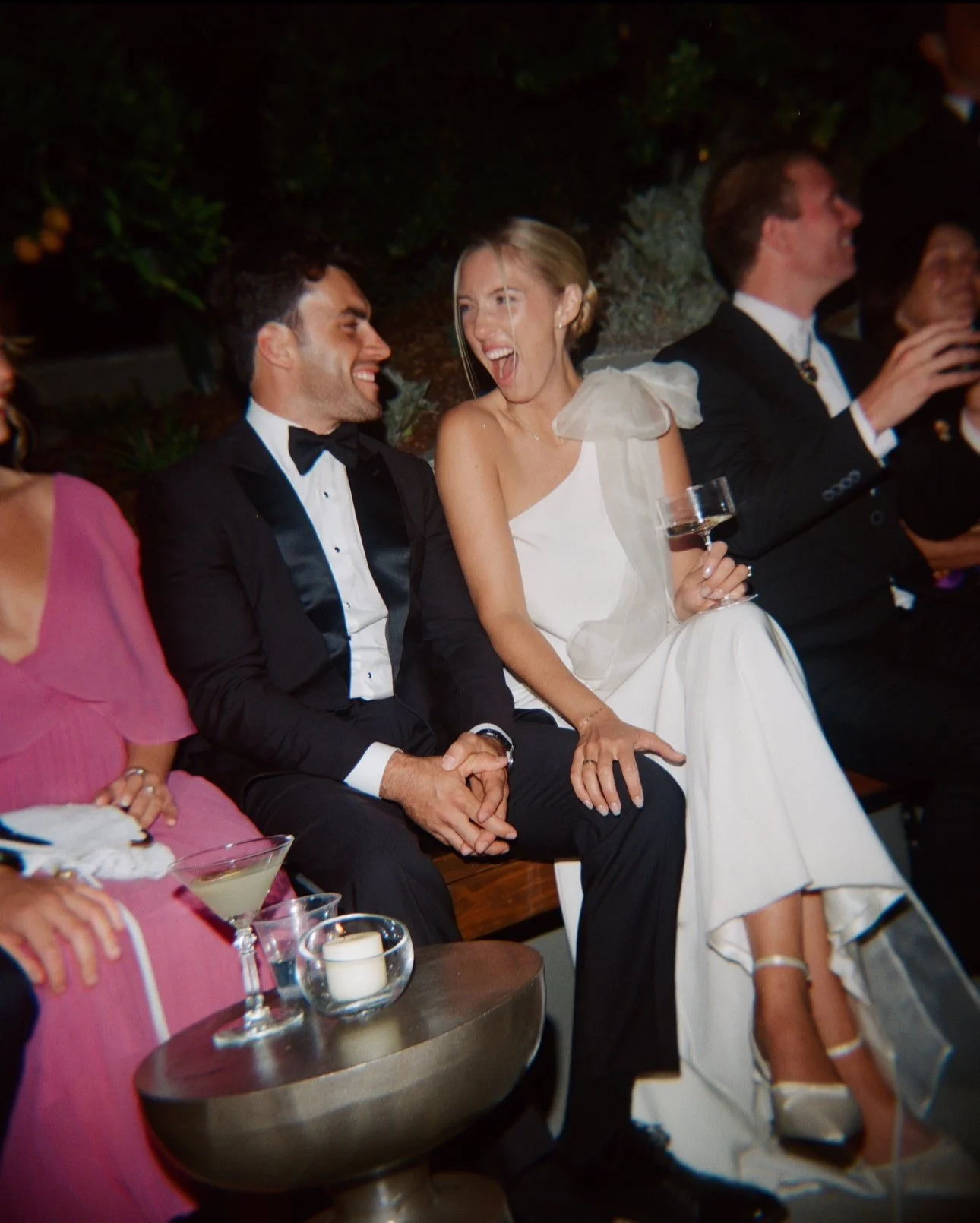 A group of people dressed in formal attire enjoying a celebration, with a man and a woman smiling and holding hands, and the woman holding a glass of wine.