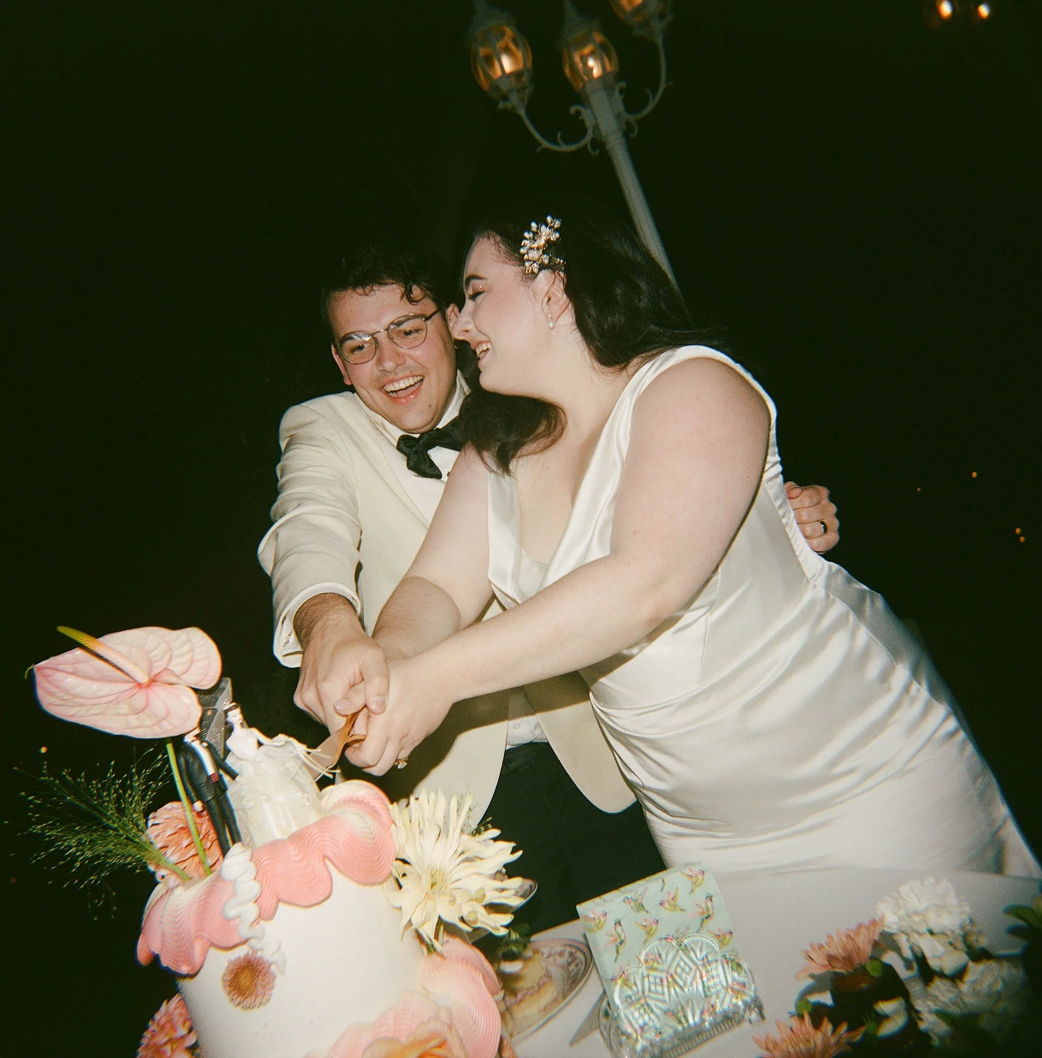 A happy couple, dressed in wedding attire, is cutting a wedding cake together at nighttime.