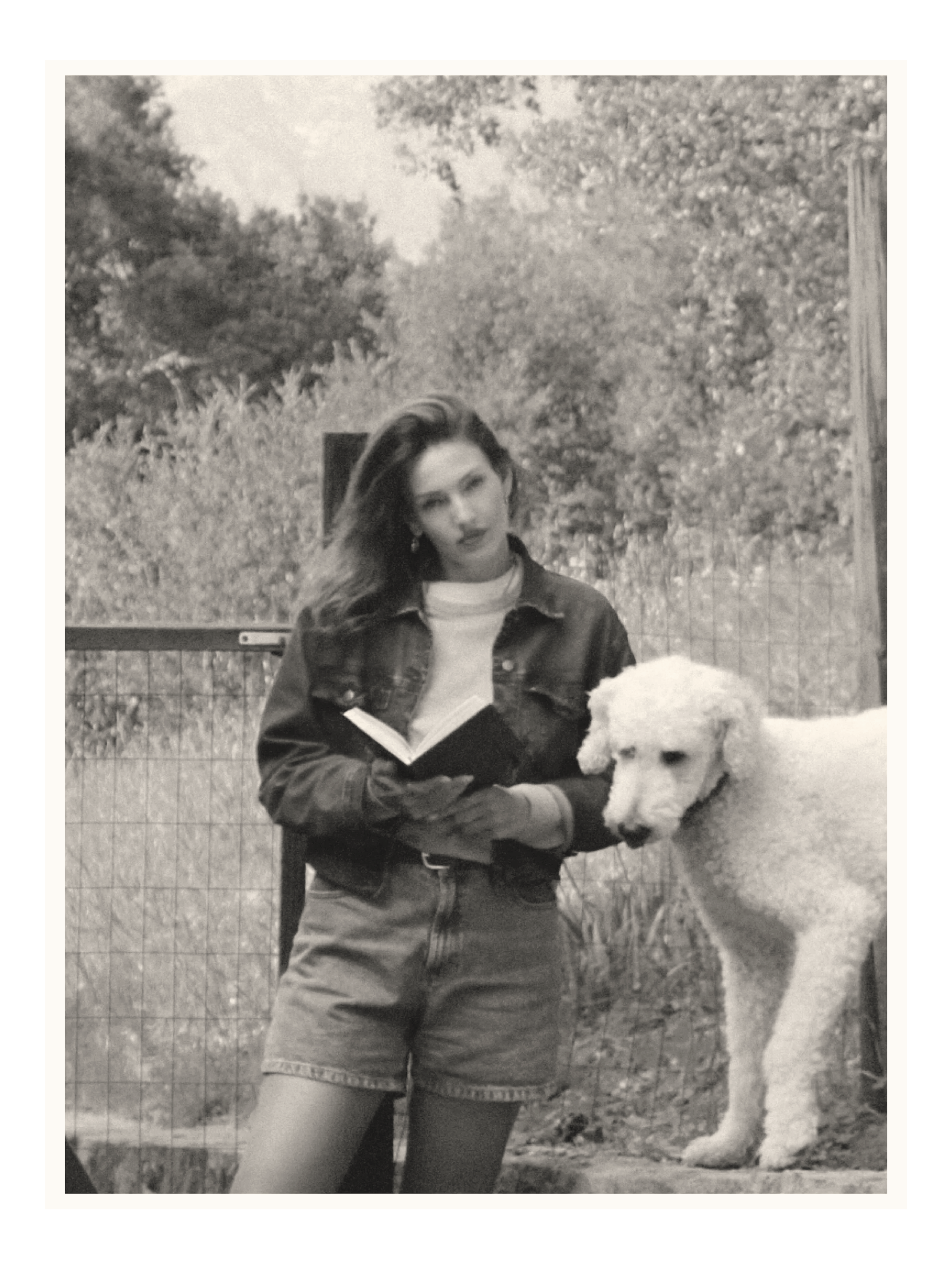 A young woman with long wavy hair, wearing a denim jacket, white shirt, and shorts, stands outdoors with a large white dog, possibly a Great Pyrenees, during daytime. She is holding a book and looking off to the side in a natural setting with trees and a fence in the background.