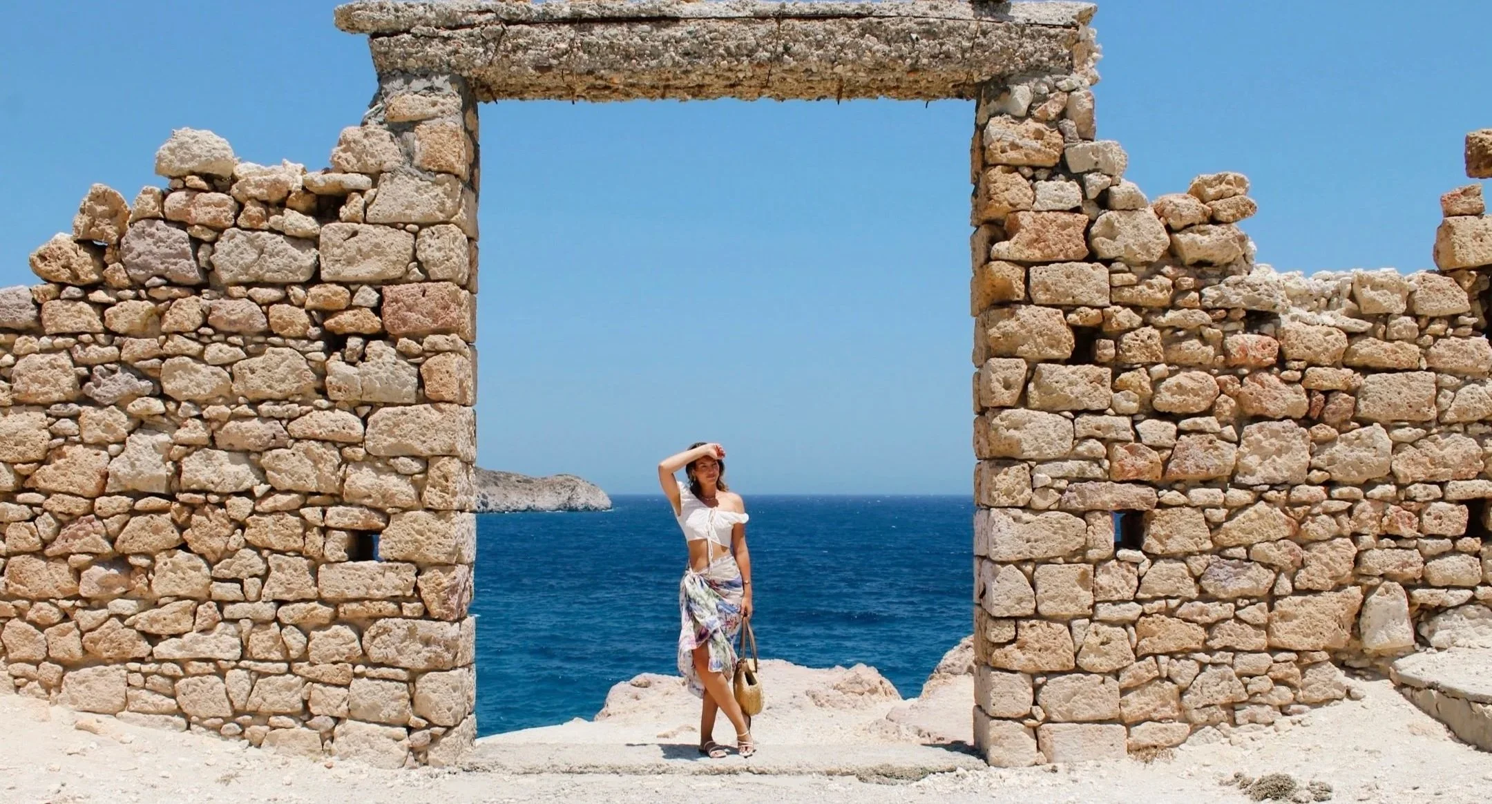 A woman in a white top and floral skirt standing beside an ancient stone archway overlooking the ocean on a sunny day.