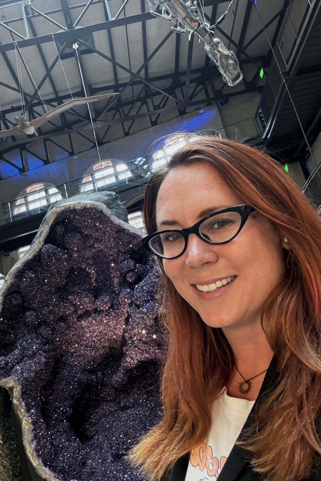A woman with red hair wearing large hoop earrings, a necklace with a pendant, and a sleeveless tropical print dress, standing on a riverside promenade at sunset with a bridge and cloudy sky in the background.
