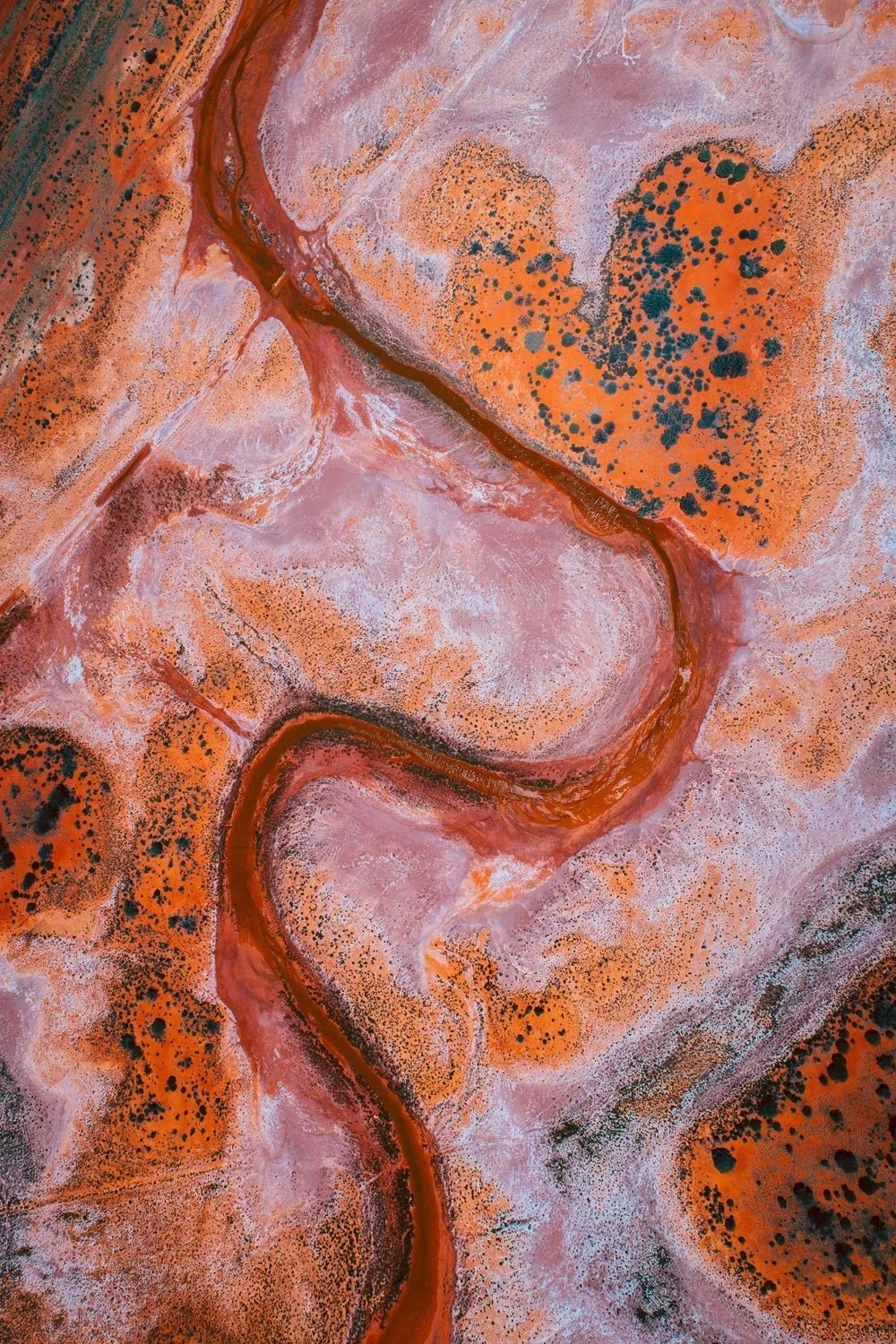 Aerial view of a winding red landscape with orange, pink, and black spots, resembling a desert or geological formation.