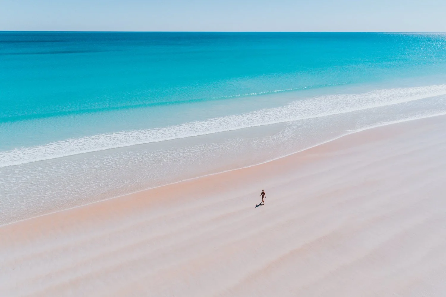A person walking alone on a wide, sandy beach with clear turquoise water and gentle waves in the background.