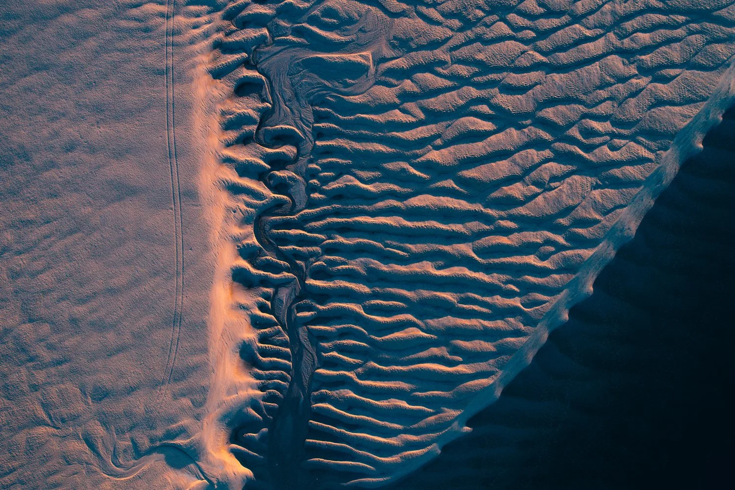 Aerial view of a sand dune with wind-formed ripples and a small winding stream, illuminated by warm sunlight.