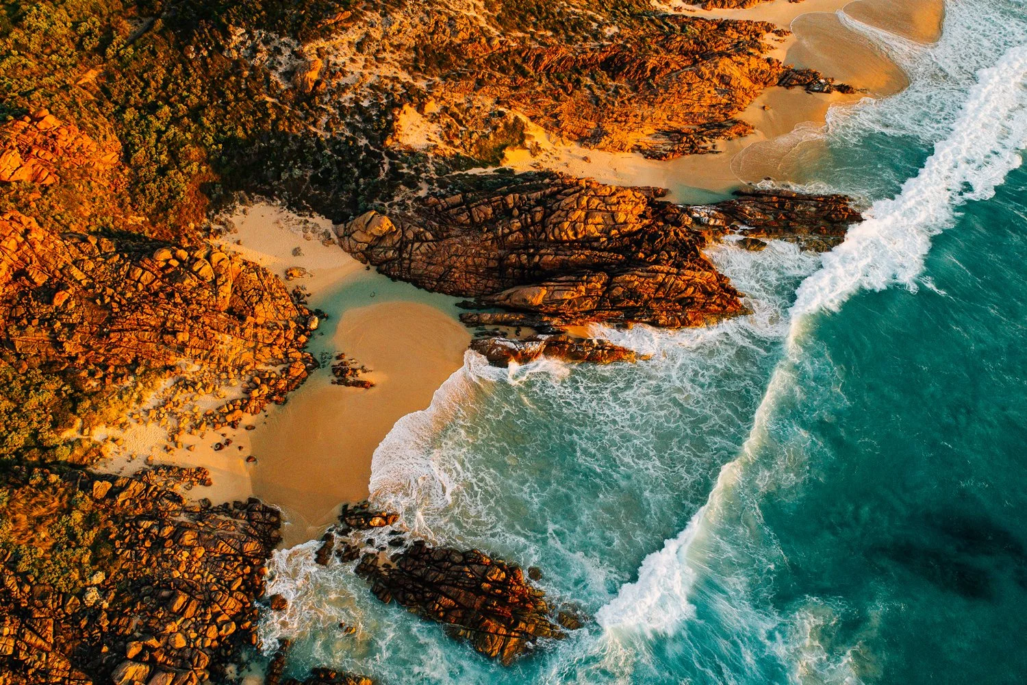 Aerial view of a rugged coastline with sandy beaches, rocky formations, and turquoise ocean waves crashing onto the shore at sunset.