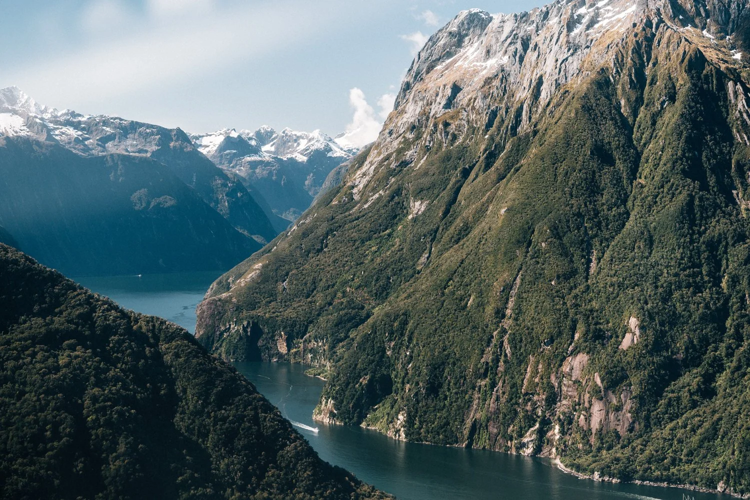 Scenic view of a fjord surrounded by tall, green, snow-capped mountains under a partly cloudy sky in Milford Sound New Zealand