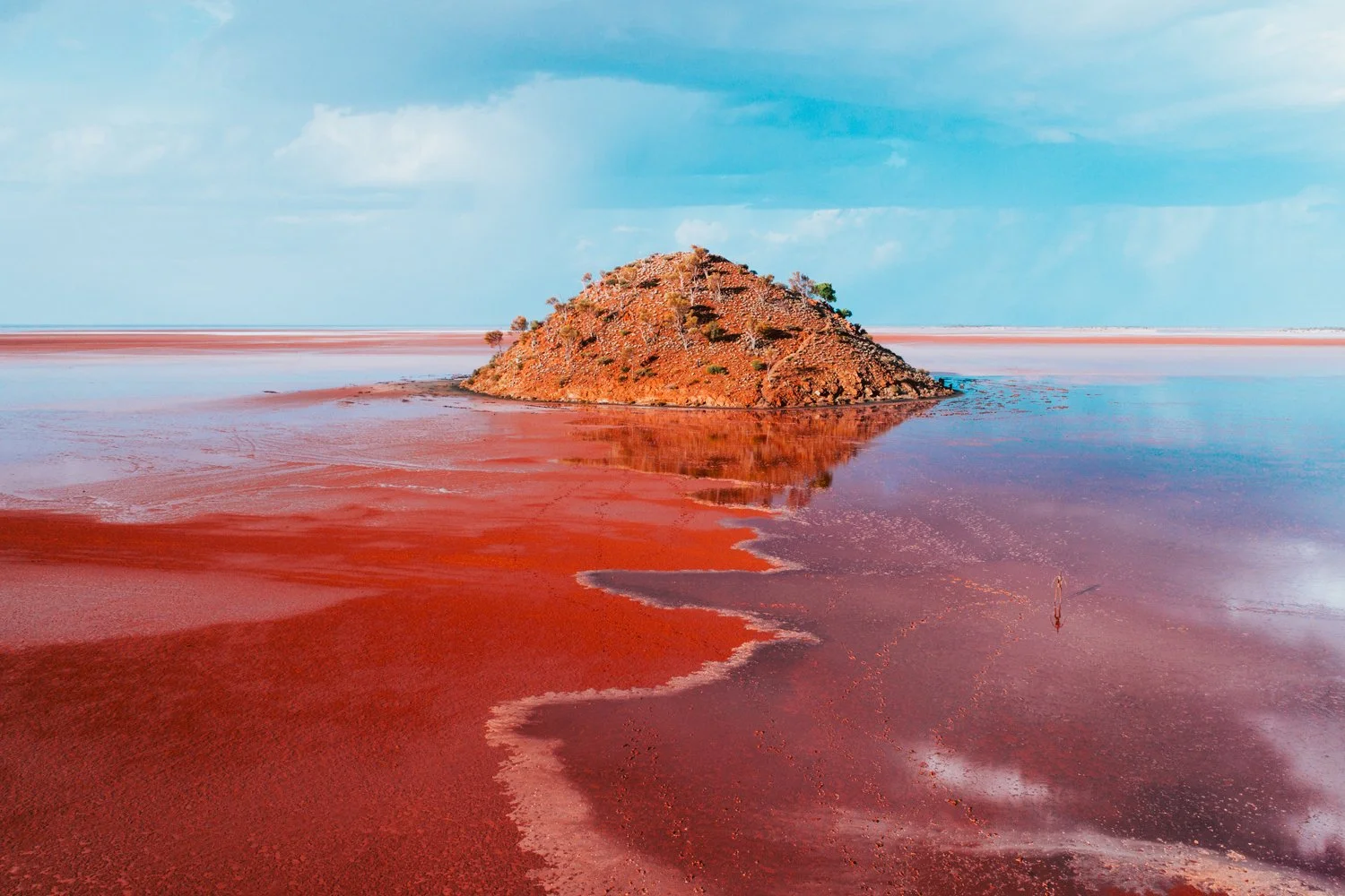 A small hill covered in shrubs rises from a red salt lake, with reflections of the hill in the water and a partly cloudy sky overhead.
