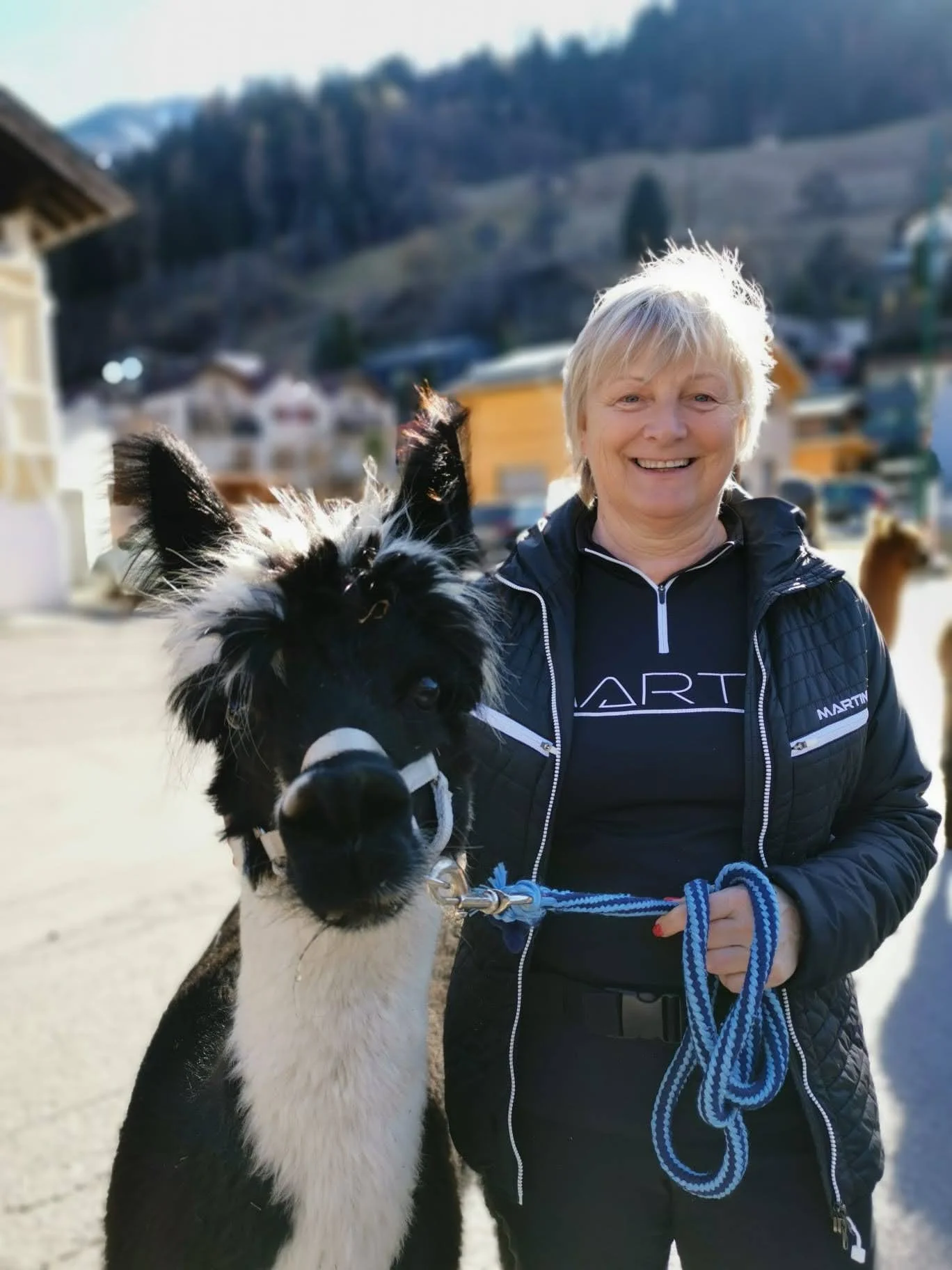Lächelnde ältere Frau hält ein schwarzes und weißes Pony an einer Leine in einer Stadt mit Bergen im Hintergrund.