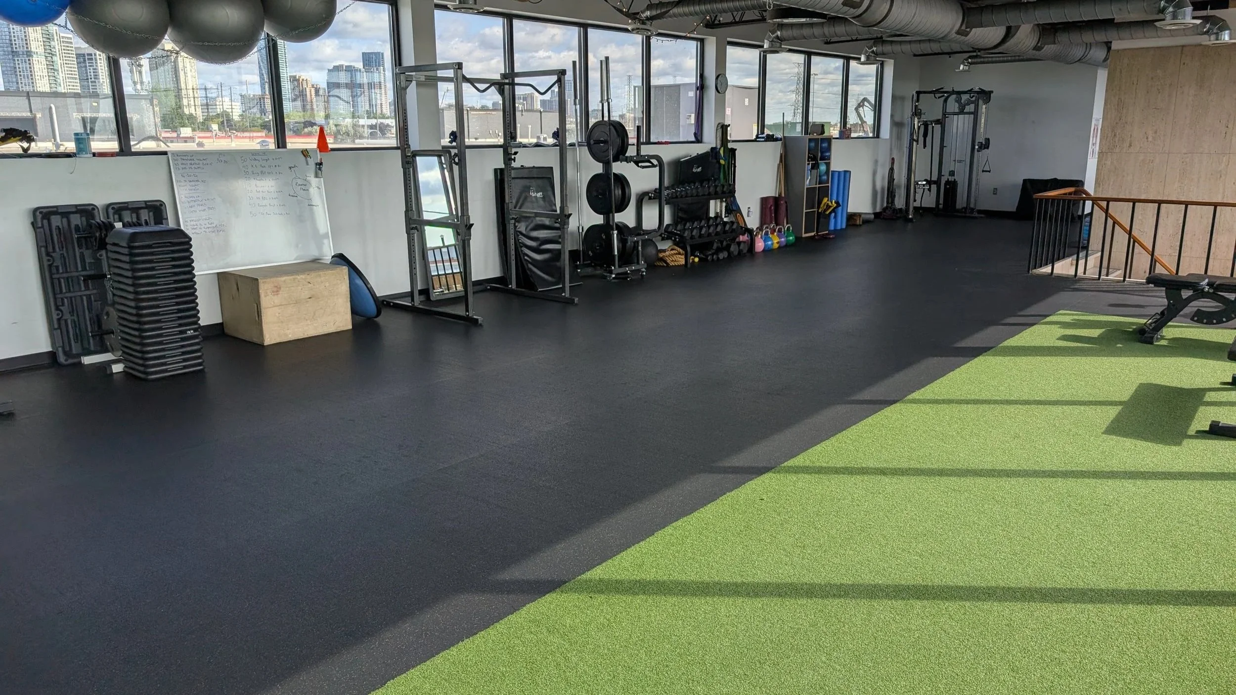 Interior of a modern gym with black rubber flooring, green turf area, and large windows showing city buildings outside. Fitness equipment includes stacked step platforms, a whiteboard, plyometric boxes, weights, kettlebells, resistance bands, and a workout bench.