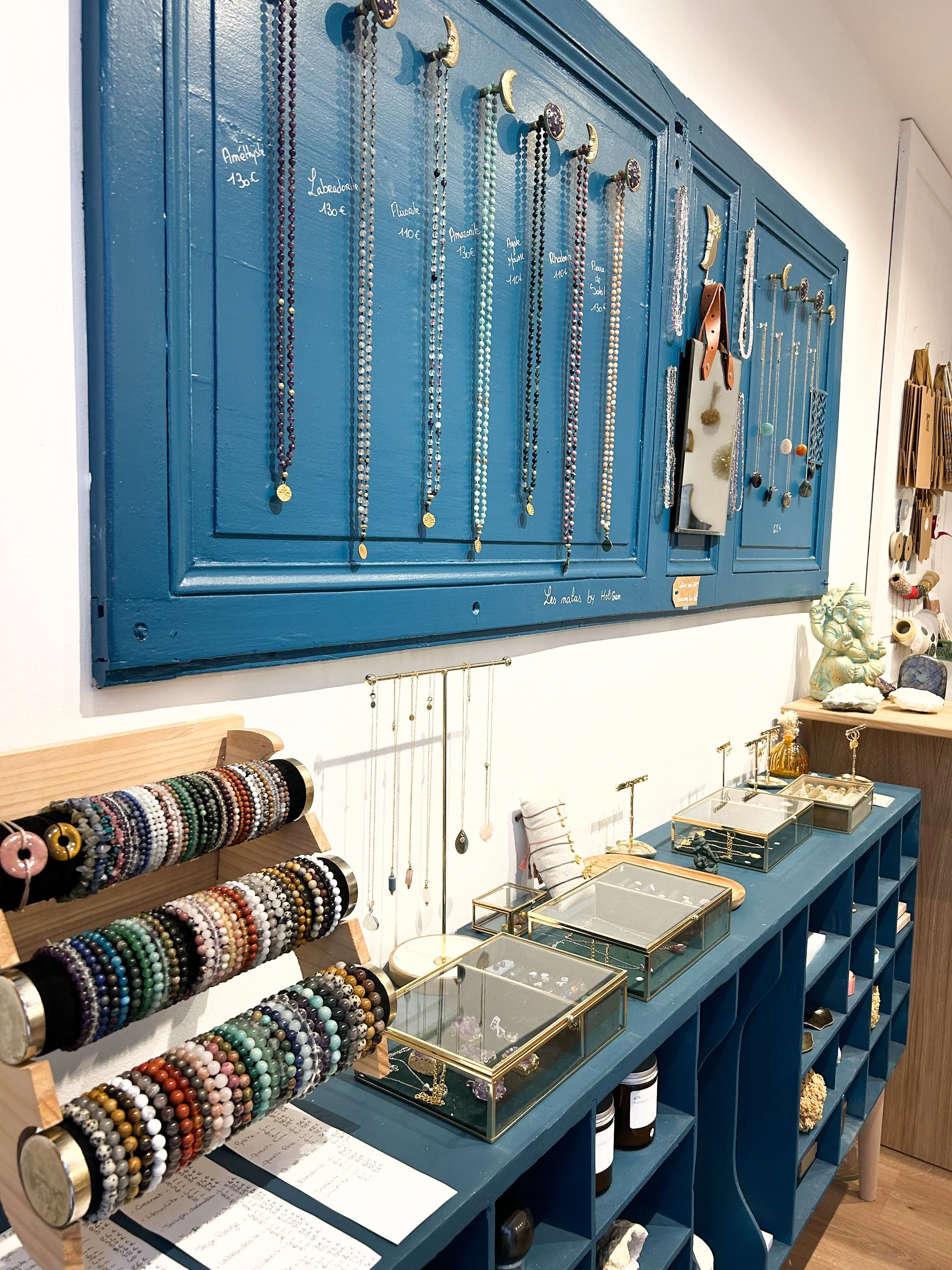 Un mur bleu avec des colliers de perles suspendus, un support à bijoux avec beaucoup de bracelets colorés, des boîtes en verre contenant des bijoux, et des figurines décoratives sur une étagère.