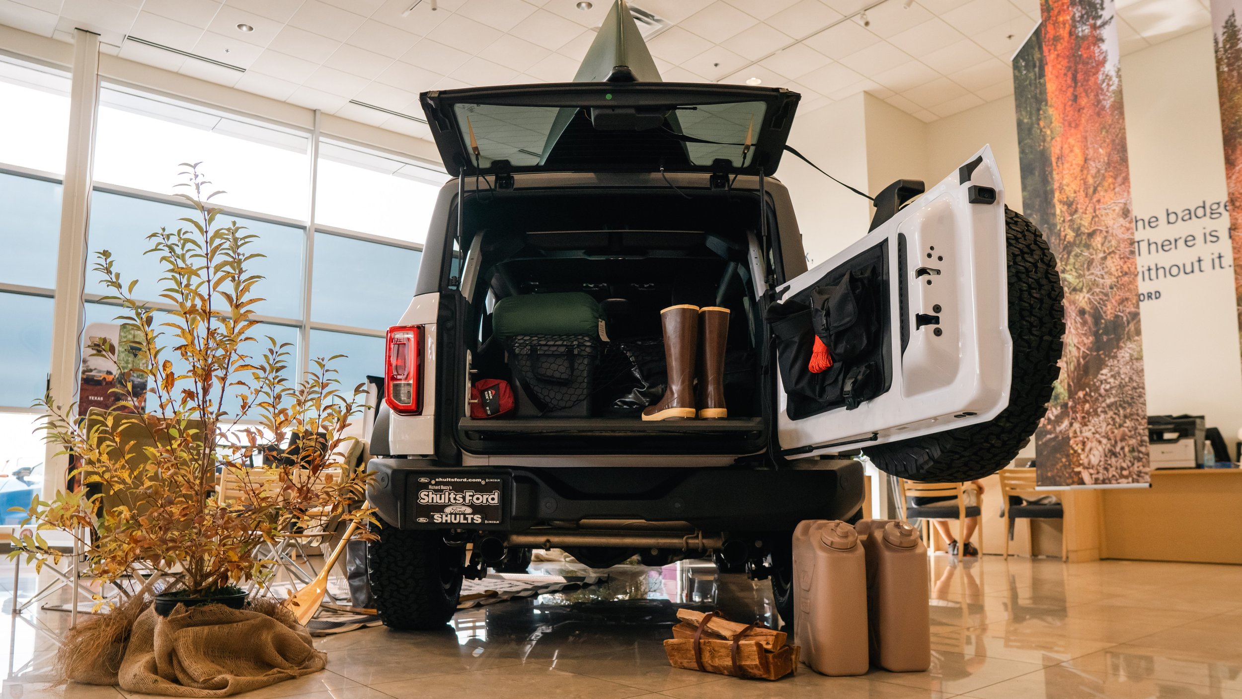 The open back of a white Jeep vehicle displayed indoors, filled with outdoor gear including a pair of rain boots, a bag, and a hat. Surrounding the vehicle are potted plants and stacked logs, likely part of an indoor showroom.