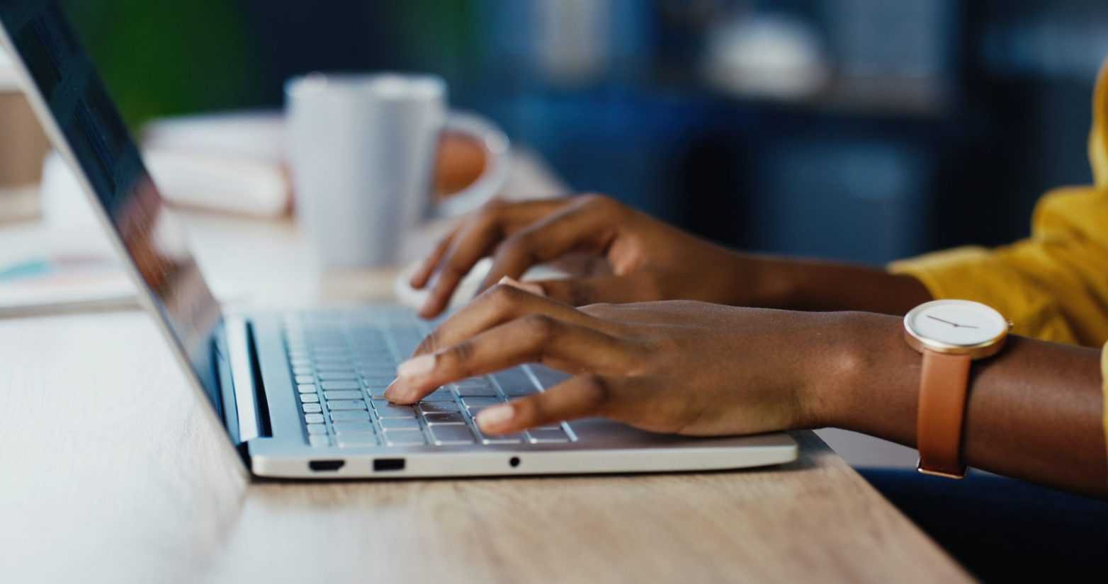 A person using a laptop on a wooden table, with a white mug in the background, wearing a wristwatch with a round face and brown strap.