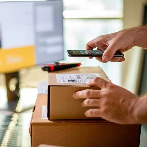 Person scanning a package with a smartphone near a computer monitor.