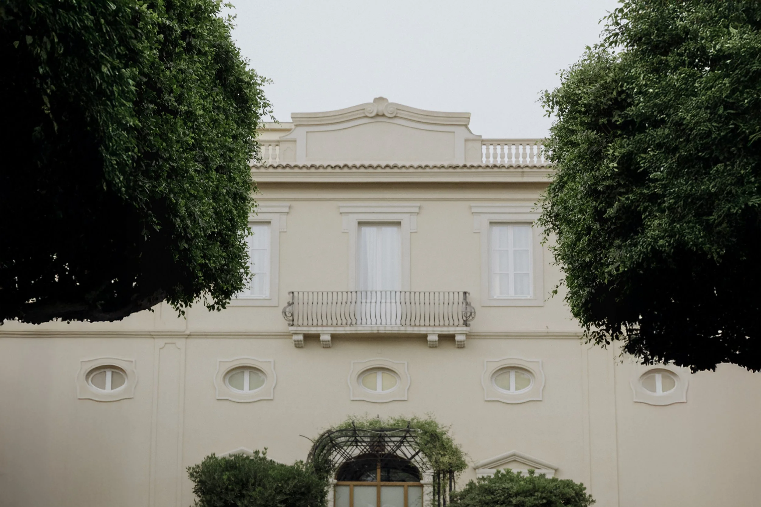 Front view of a white building with a balcony, arched window, surrounded by green trees.