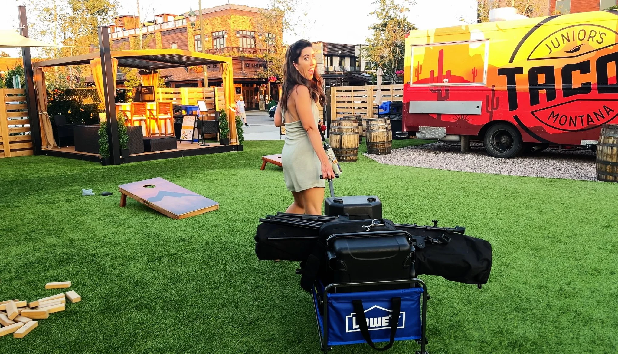 A woman with long dark hair, wearing a light-colored dress, stands outdoors on artificial green grass holding a suitcase. There is game equipment on the ground, including a cornhole board and wooden blocks. In the background, there are wooden barrels, a food truck with