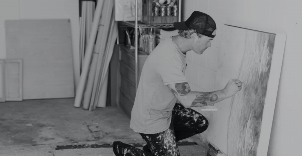 A young man with tattoos, wearing a backwards cap, painting on a large canvas in an art studio, with art supplies and rolled papers in the background.